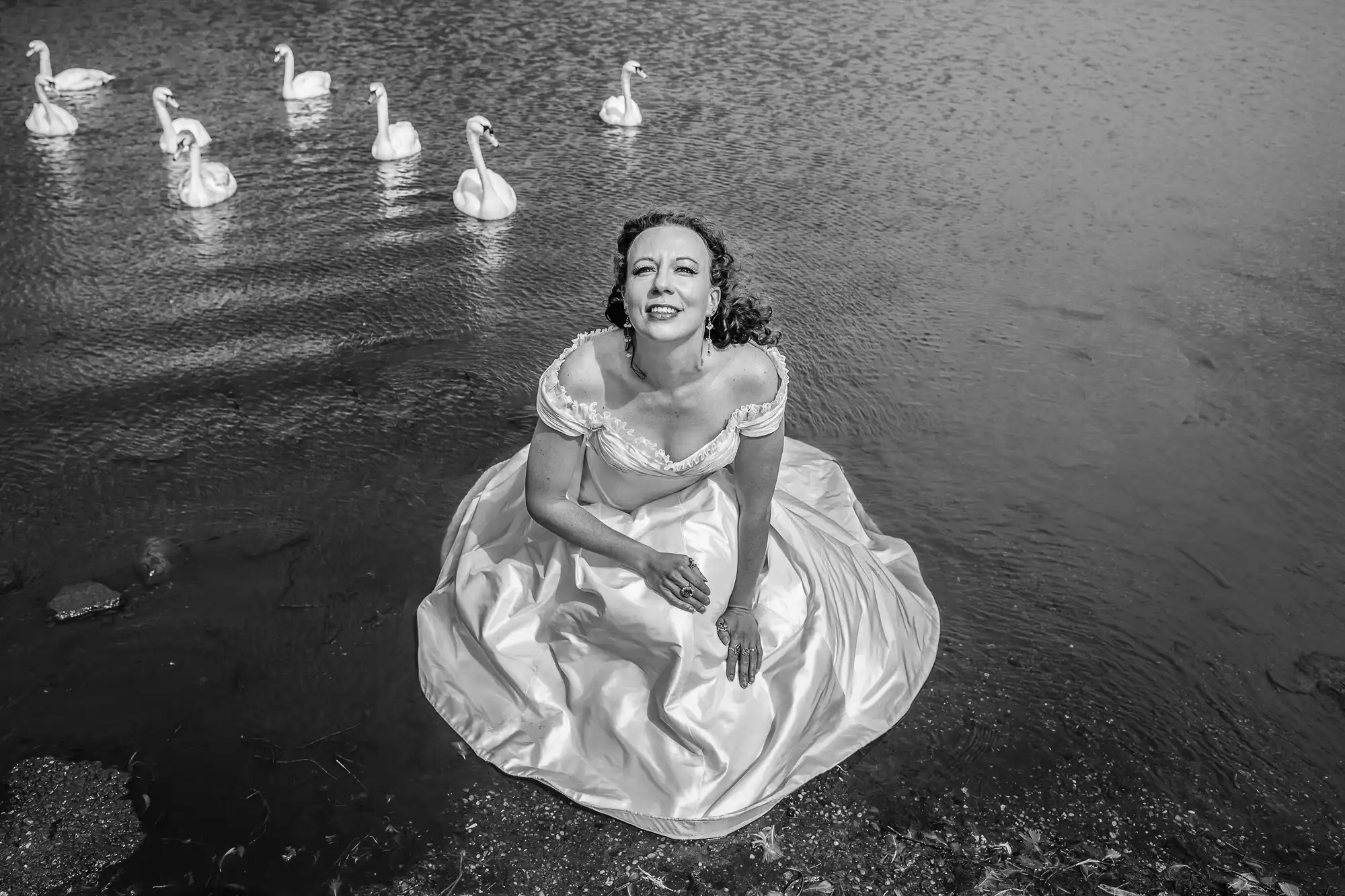 Woman in an elegant white dress sitting in shallow water surrounded by swans, black and white photography.