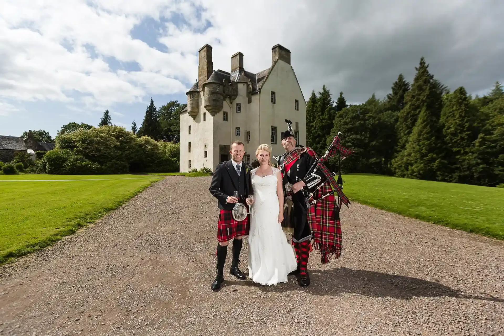 A bride and groom stand with a bagpiper, Pipe Major Iain Grant, in front of historic Tullibole Castle on a sunny day, all dressed in traditional Scottish attire.