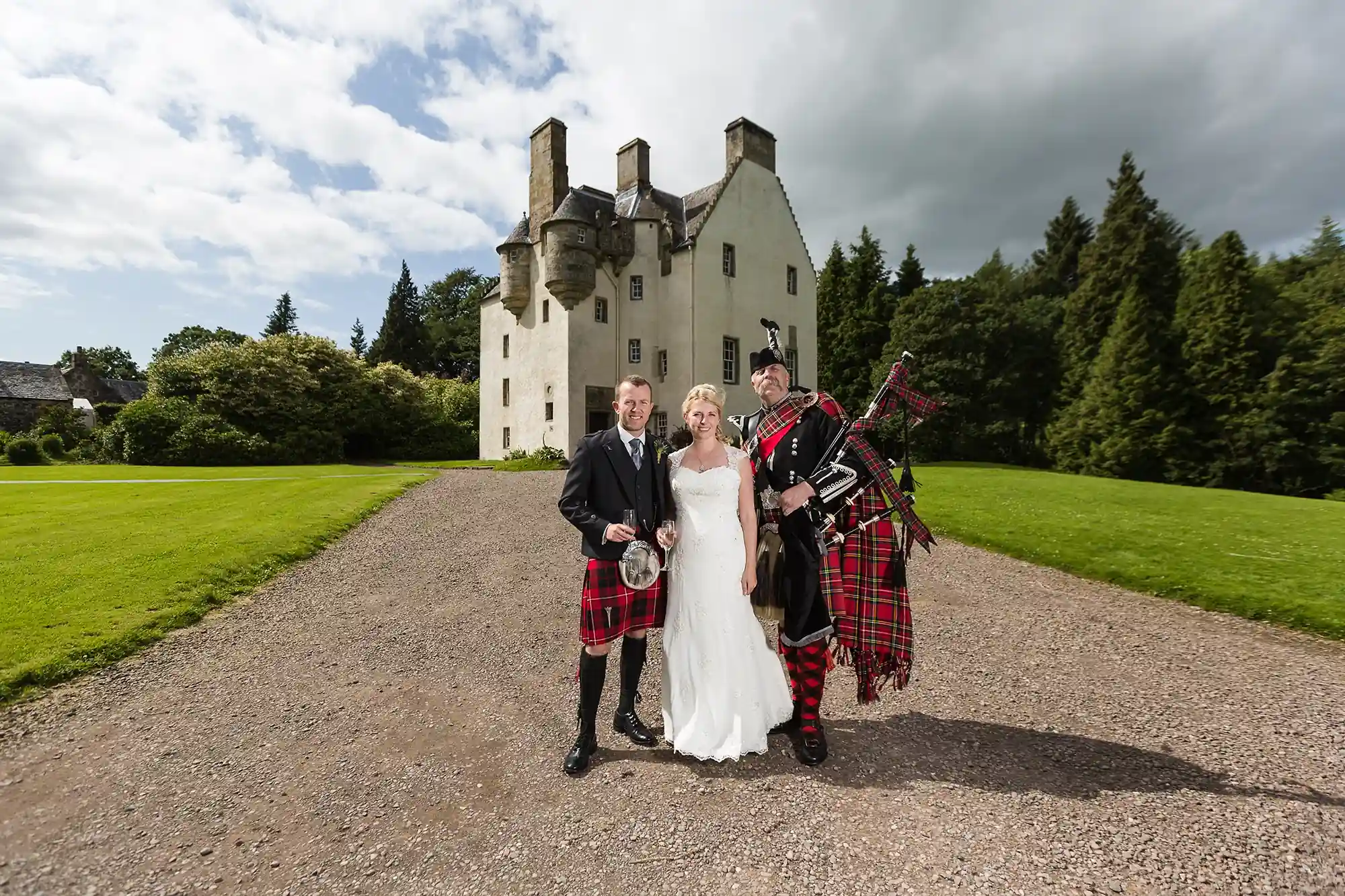 A bride and groom stand with a bagpiper in front of a historic castle on a sunny day, all dressed in traditional scottish attire.