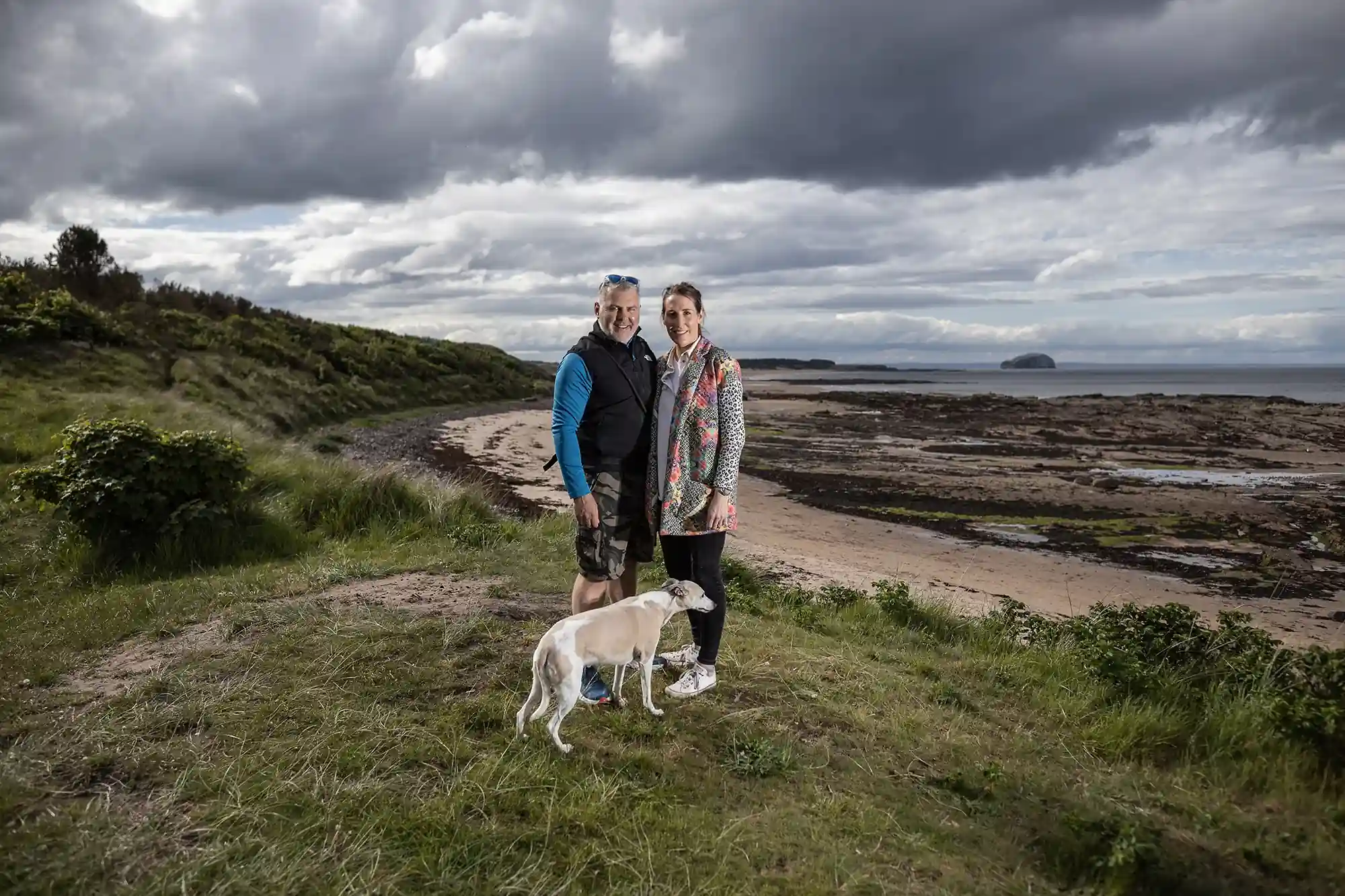 A couple with a dog standing by a coastal pathway under a cloudy sky, with the ocean and a distant island in the background.