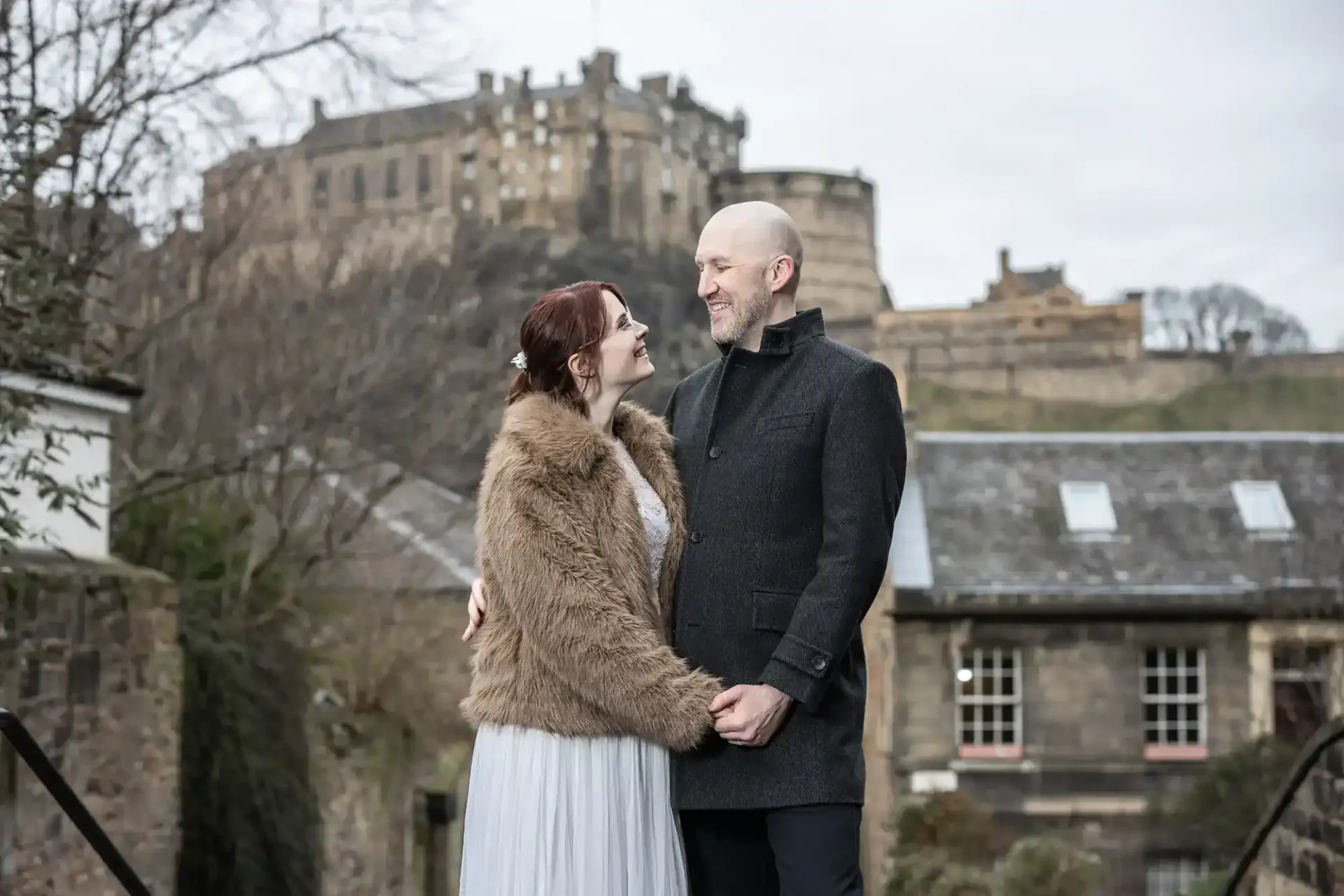 A couple stands outdoors in front of a historic stone building, holding hands and smiling at each other on a cloudy day.