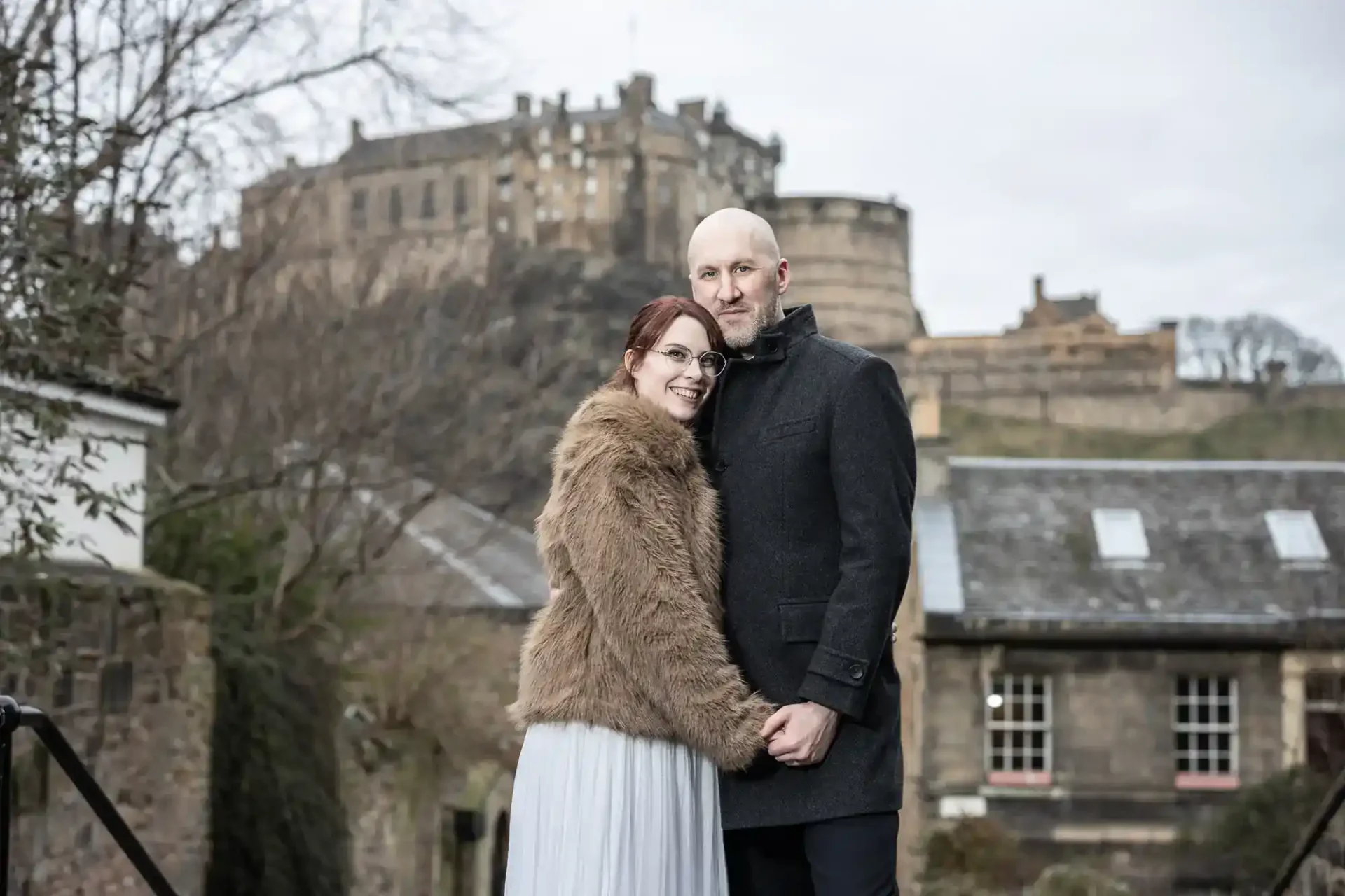 A couple stands close together, holding hands and posing outdoors with a large historic castle on a hill in the background.