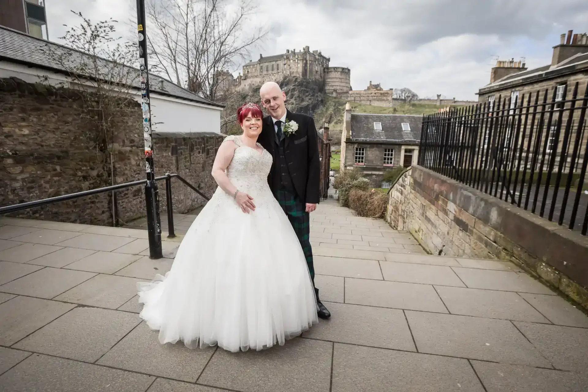A bride in a white gown and a groom in a kilt pose together on a stone path, with Edinburgh Castle visible in the background.