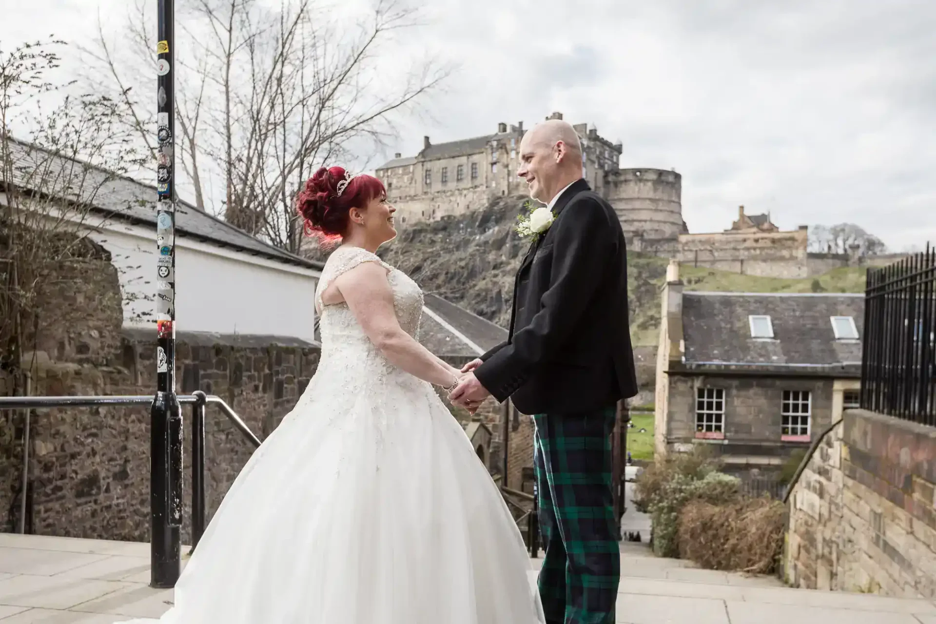 A bride and groom hold hands and smile at each other outdoors, with a castle on a hill in the background.