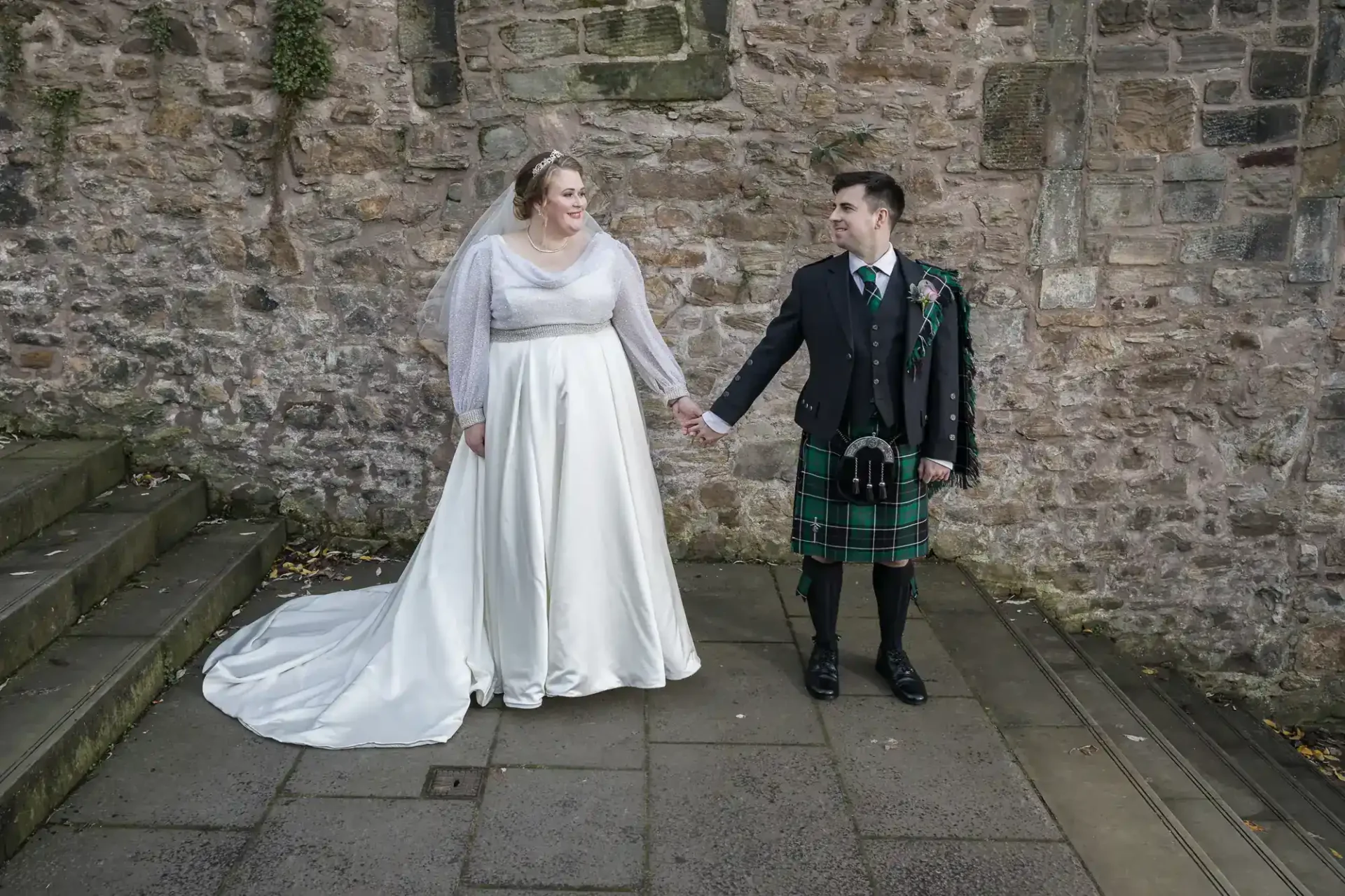 A bride in a white gown and a groom in a black jacket and green tartan kilt stand holding hands in front of a stone wall.