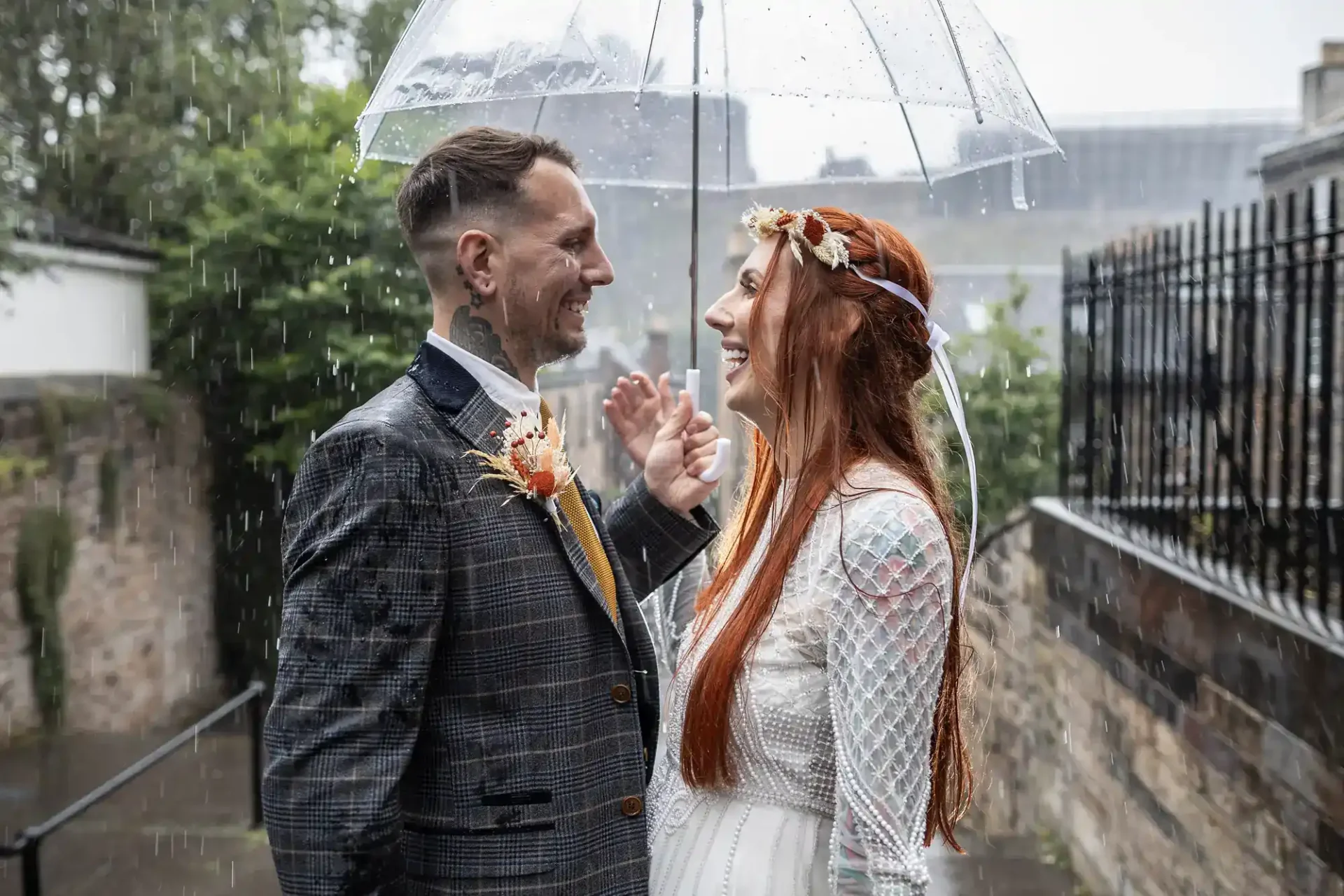 A bride and groom stand under a clear umbrella in the rain, smiling at each other outdoors on their wedding day.
