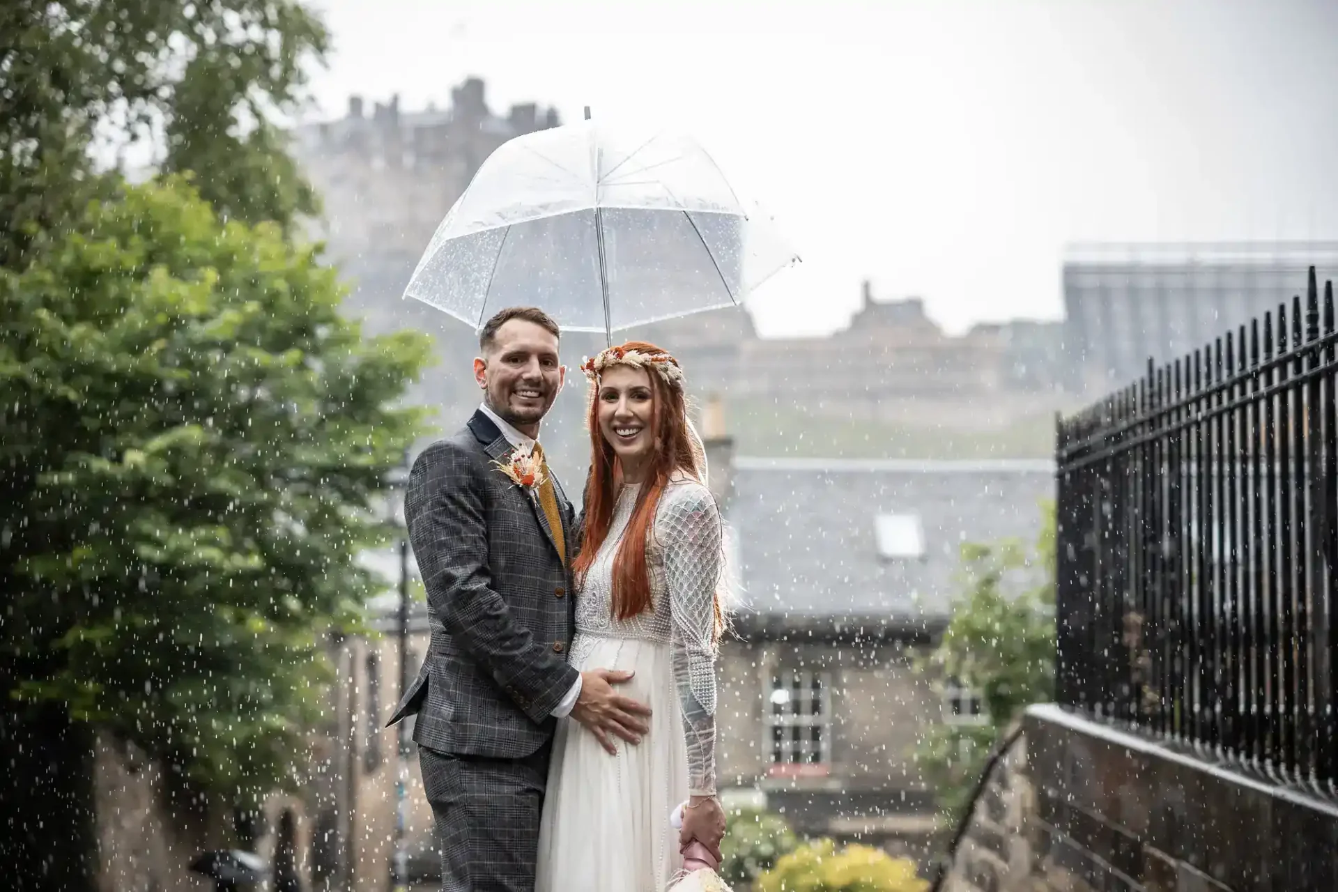 A bride and groom stand closely under a clear umbrella in the rain, smiling, with historic stone buildings and greenery in the background.