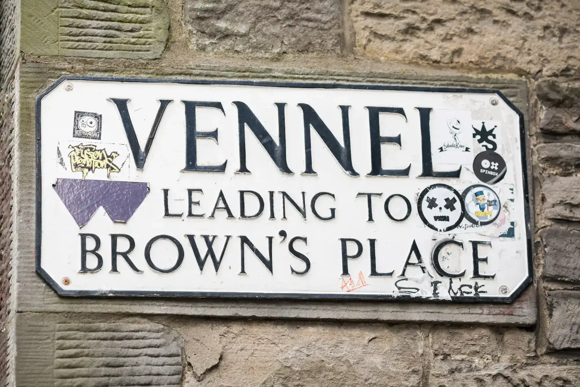 A street sign reads "Vennel Leading to Brown’s Place," partially covered with various stickers and graffiti, mounted on a stone wall.