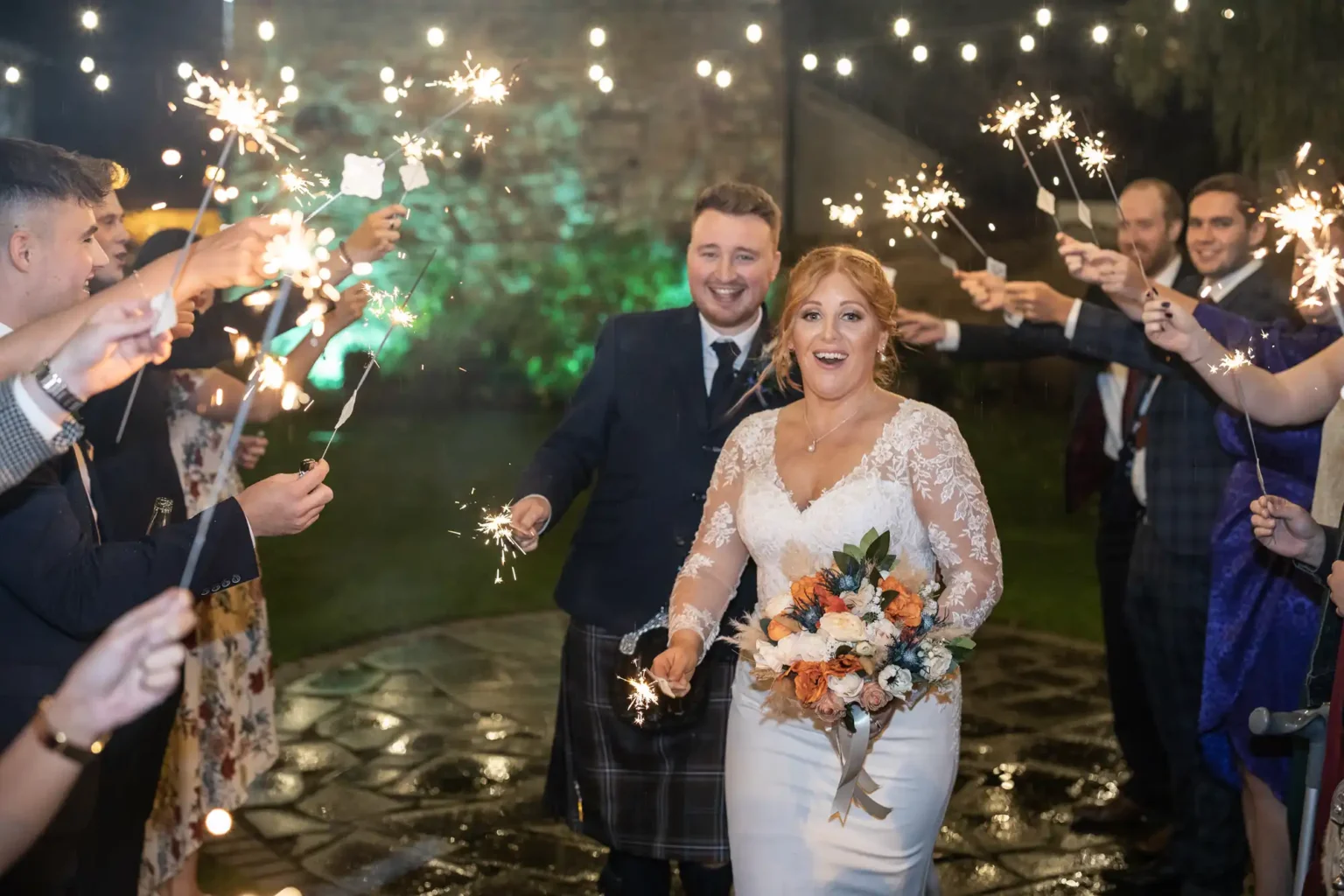 A bride and groom walk outdoors at night, surrounded by guests holding sparklers. The bride holds a bouquet and smiles, while the groom follows behind her, also smiling.