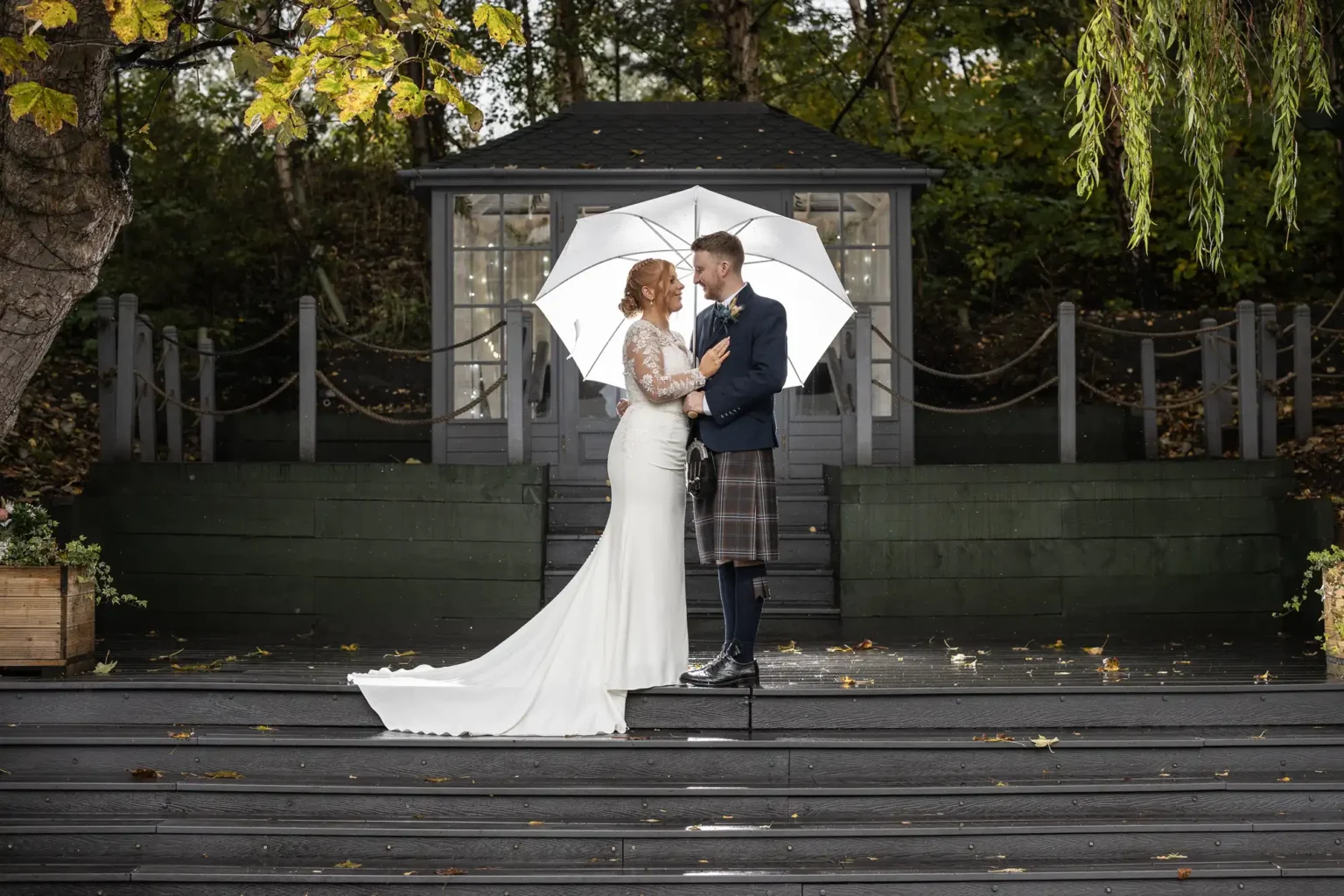 A bride and groom stand on outdoor steps under a white umbrella, facing each other, with a garden structure and trees in the background.