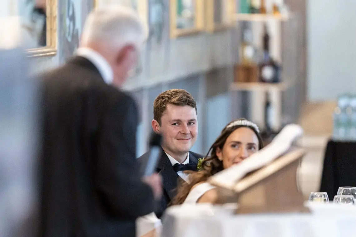 Groom smiling as he watches father of the bride deliver wedding speech
