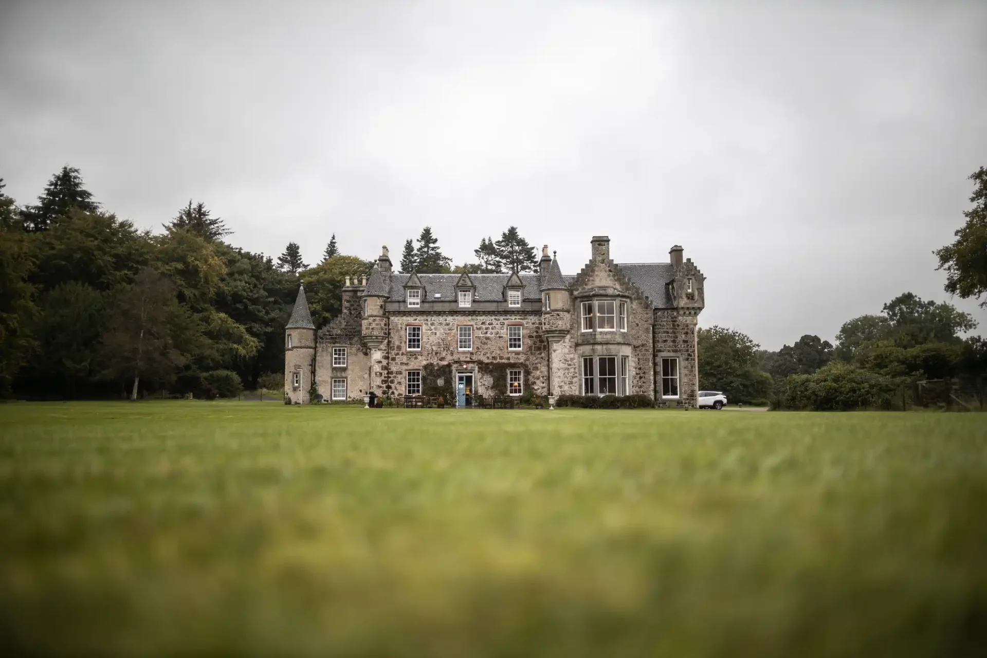 Stone mansion with multiple chimneys and turrets, surrounded by grass and trees under a cloudy sky.