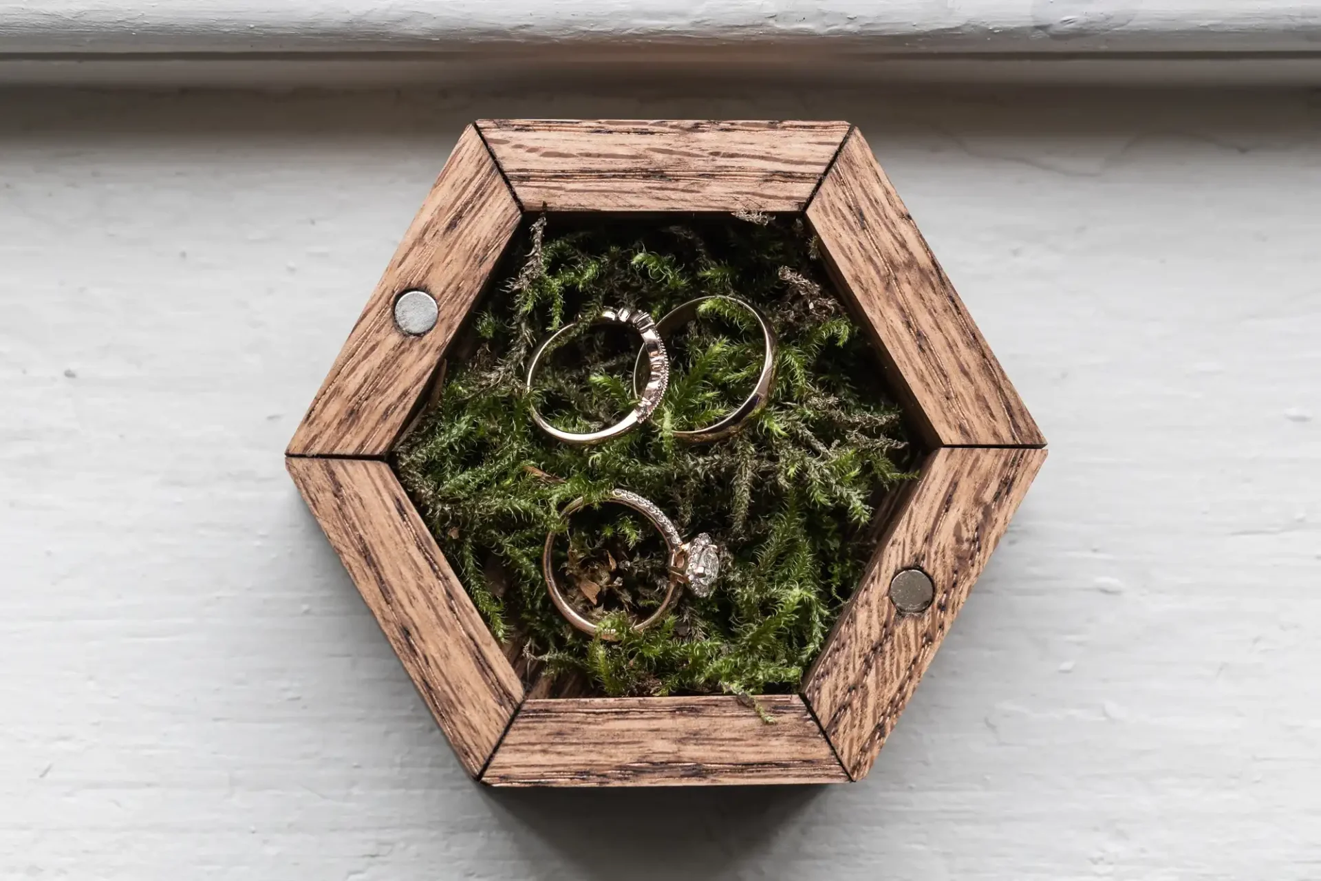 Three gold rings with diamonds are displayed on green moss inside a small hexagonal wooden box.