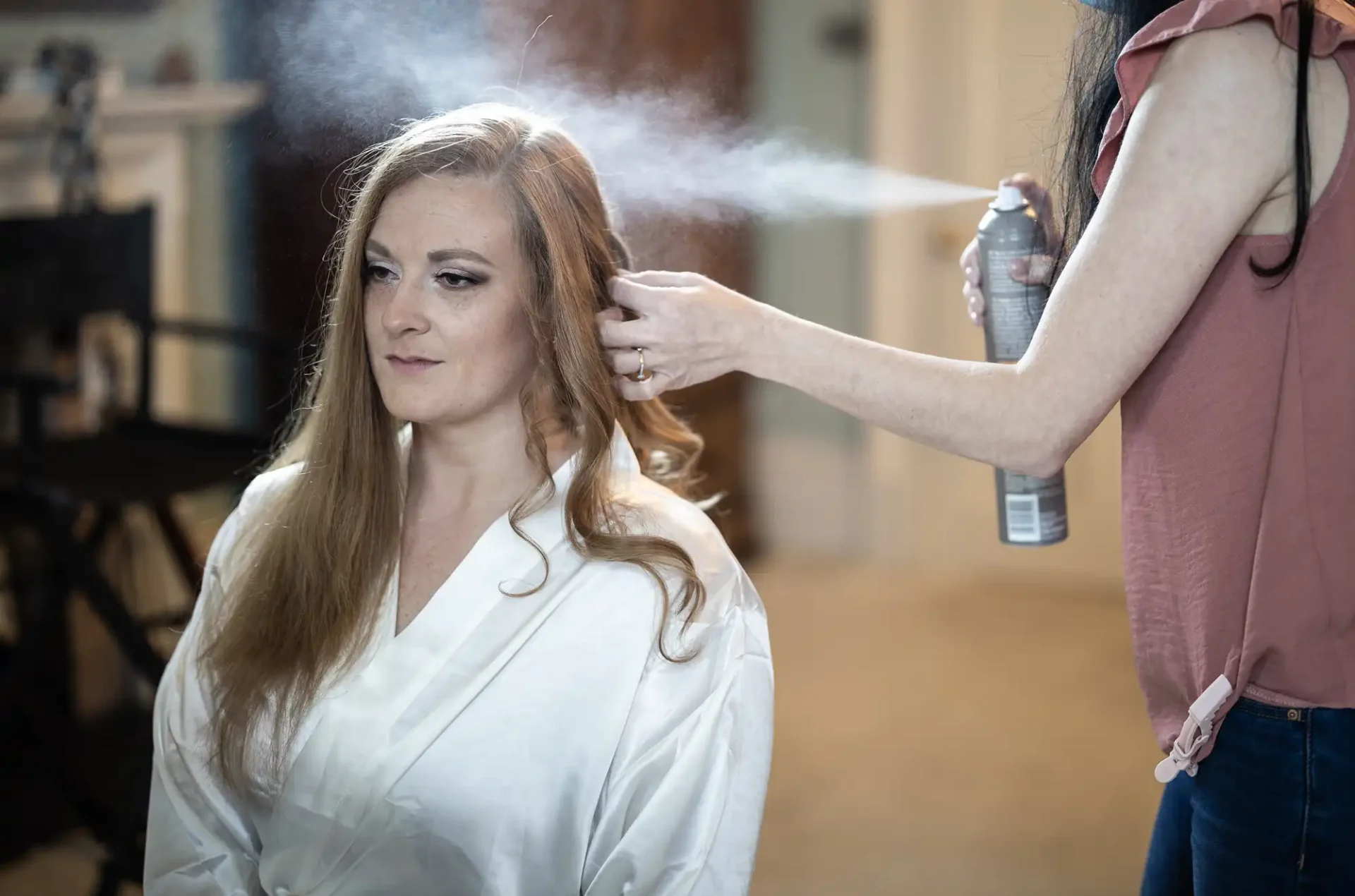 A woman in a white robe sits while another person sprays her hair with a can of hair product.