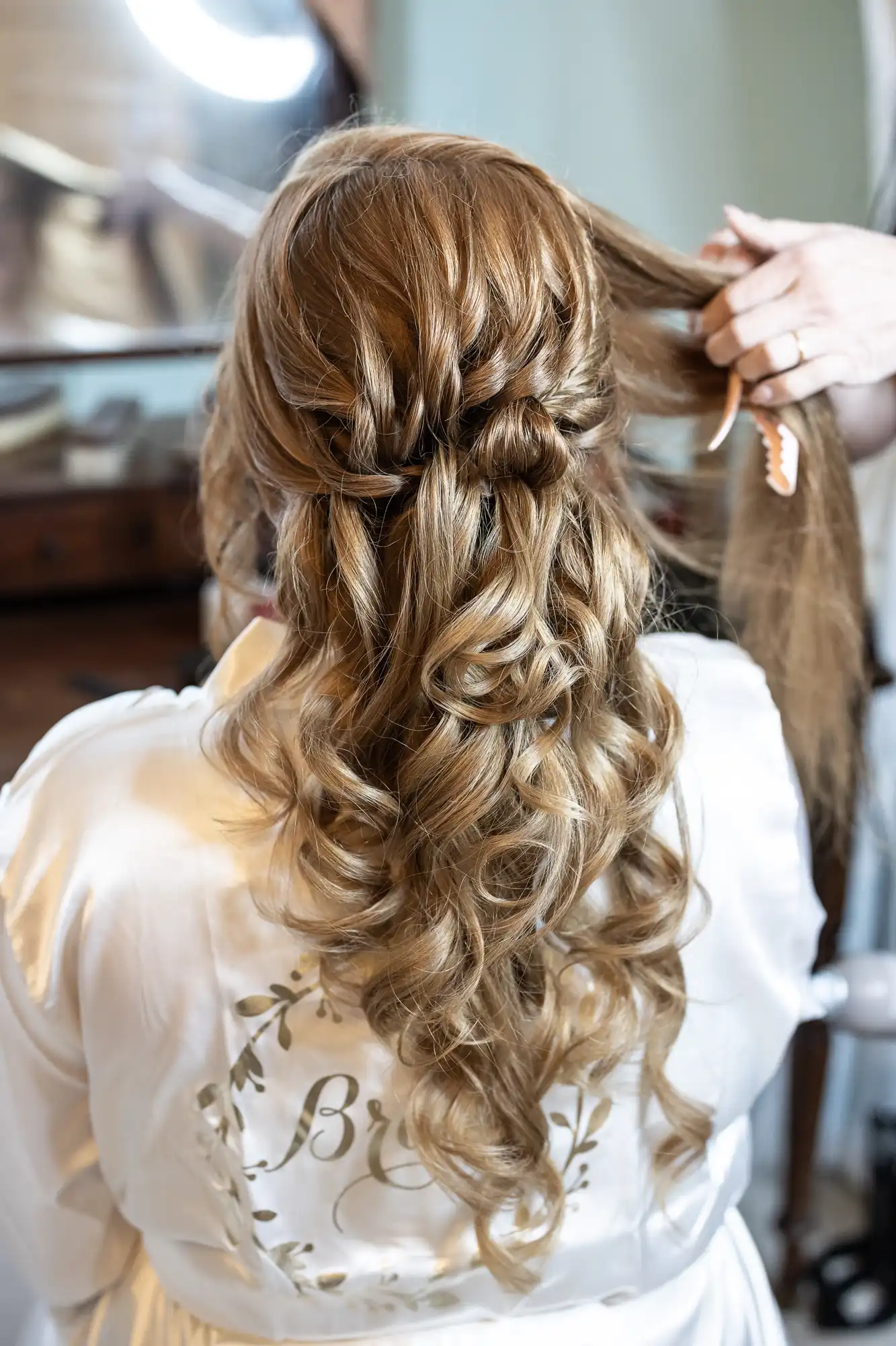 A person with long, curled brown hair styled in an intricate braid sits facing away. They wear a white robe with “Bride” written on the back.