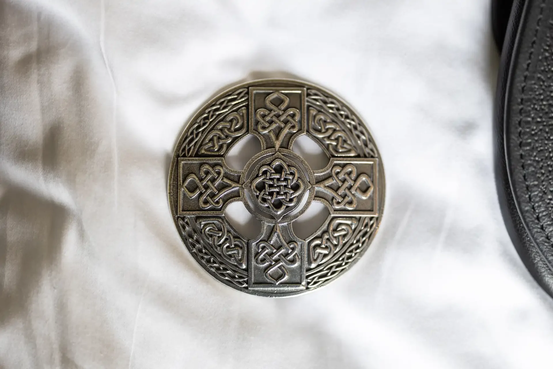 A metal Celtic knot design on a white fabric background, partially overlapping a black textured object.