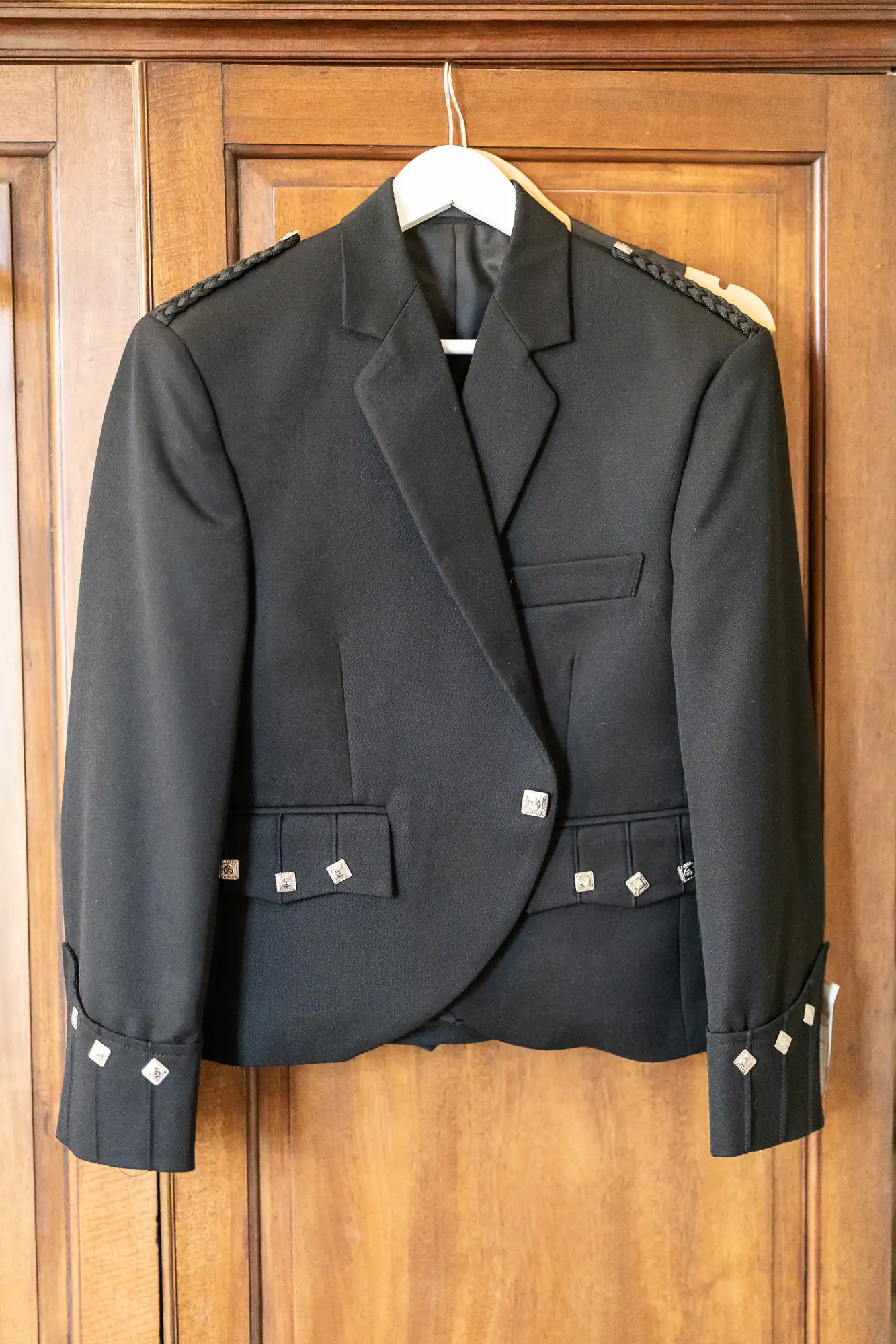 Black formal jacket with silver buttons and decorative cuff details, hanging on a wooden closet door.