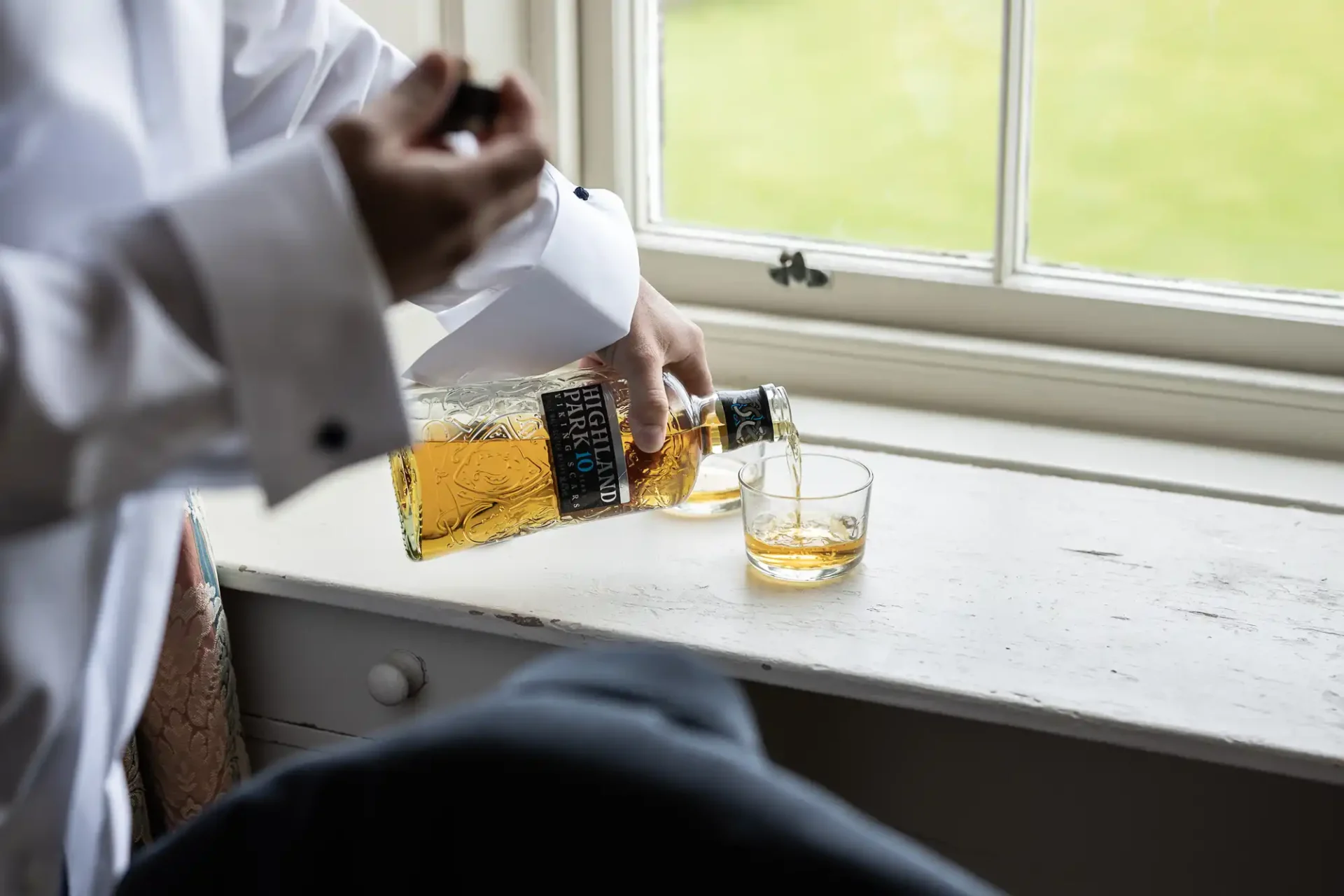 A person pours whiskey from a bottle into a glass on a windowsill.