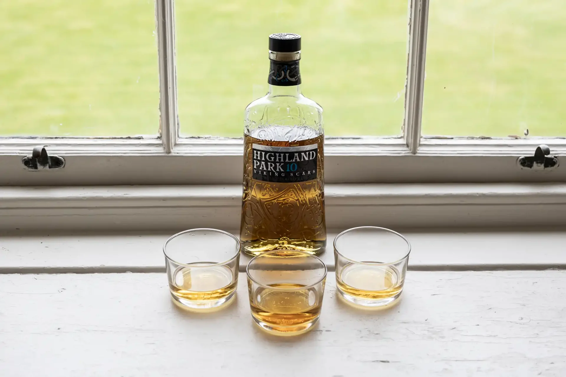 A bottle of Highland Park 10-Year-Old whisky is placed on a windowsill, accompanied by three glasses, each partially filled with the whisky.