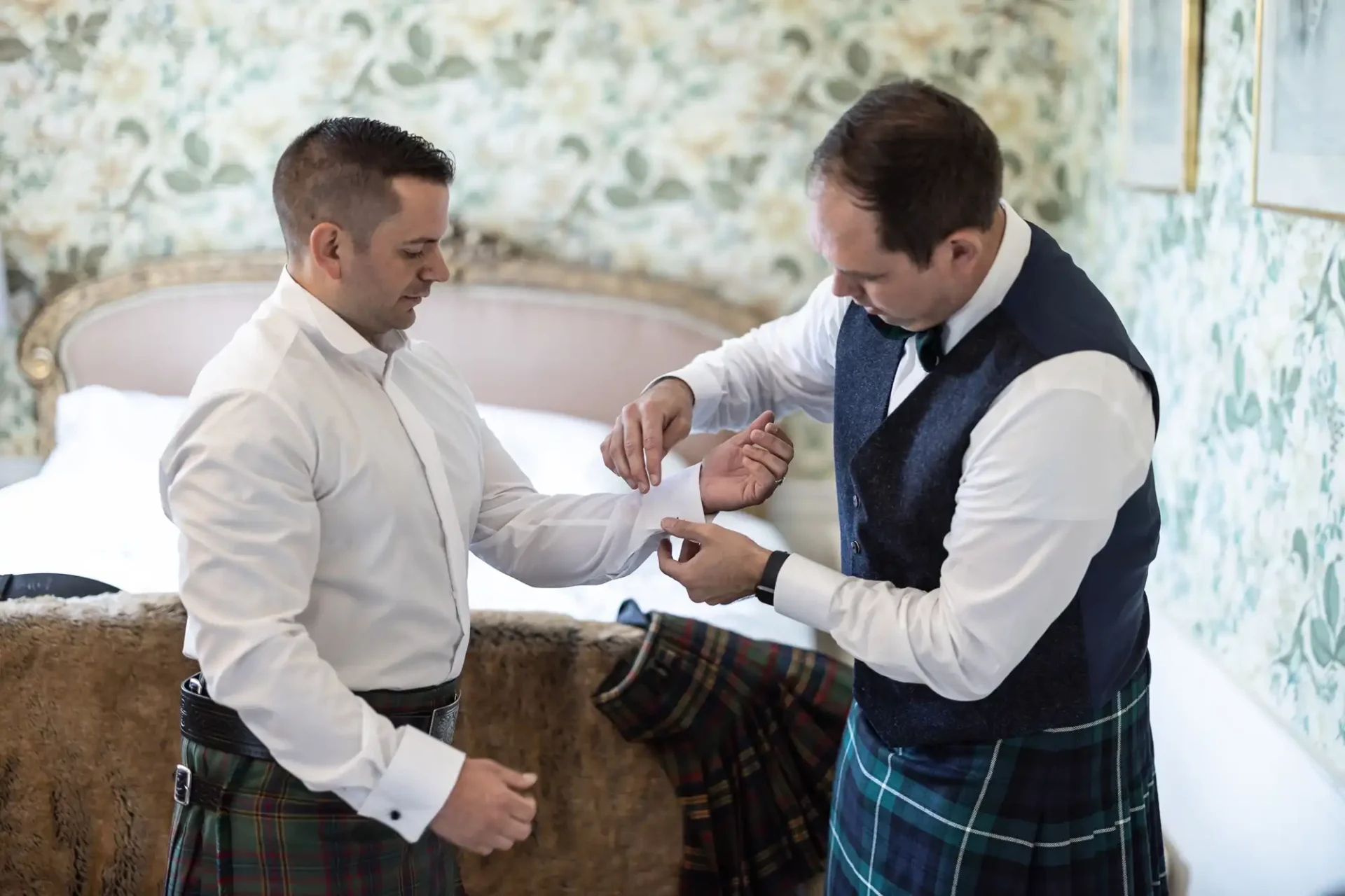 Two men in formal attire, wearing kilts, adjust a cufflink in a bedroom with floral wallpaper.