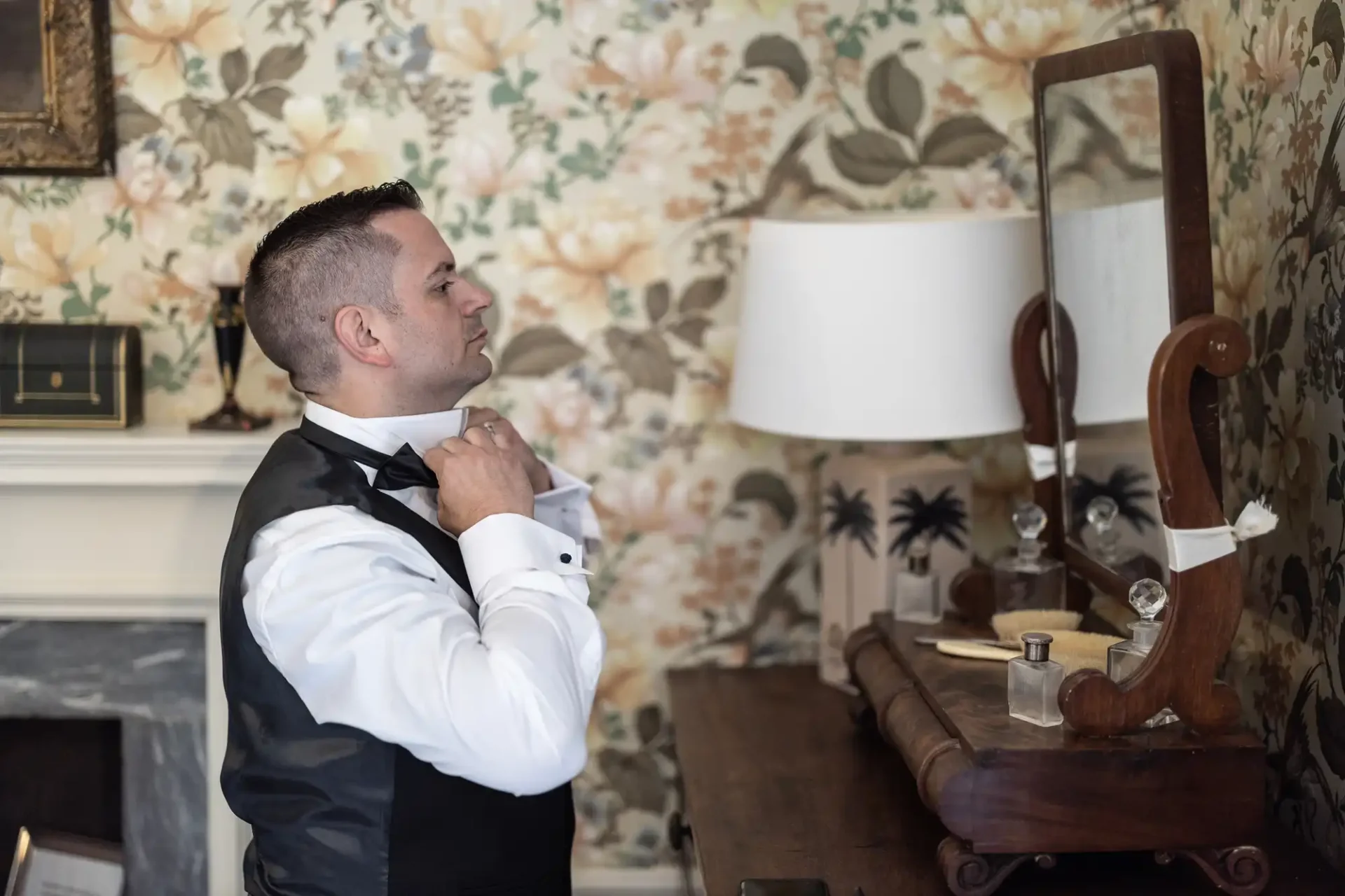 A man in formal attire adjusts his bow tie in front of a mirror in a room with floral wallpaper and a wooden dresser.