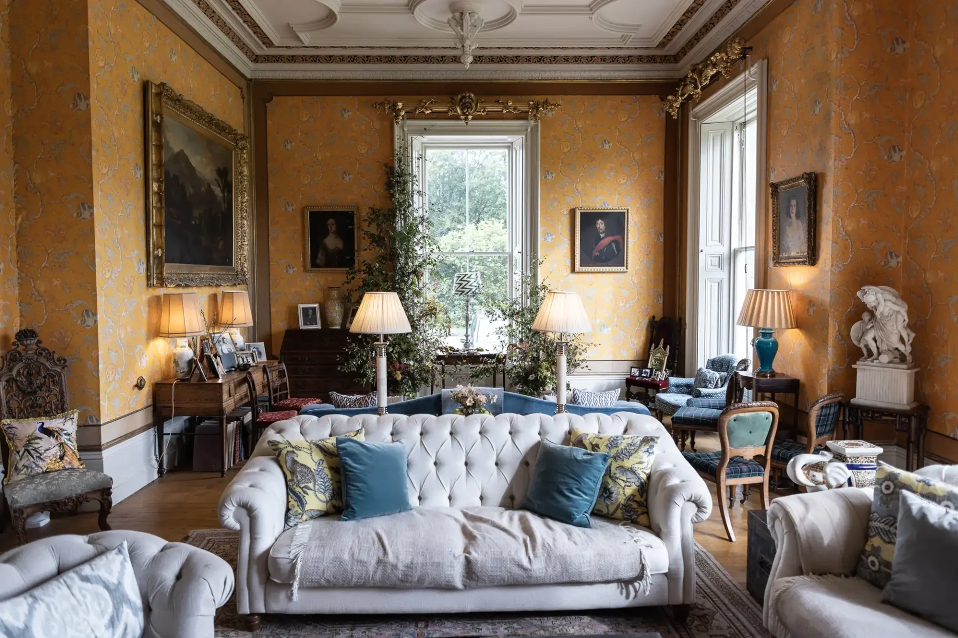 Elegant living room with yellow wallpaper, large windows, and ornate ceiling. Features white sofas, blue cushions, lamps, paintings, and a small table with various decorative items.