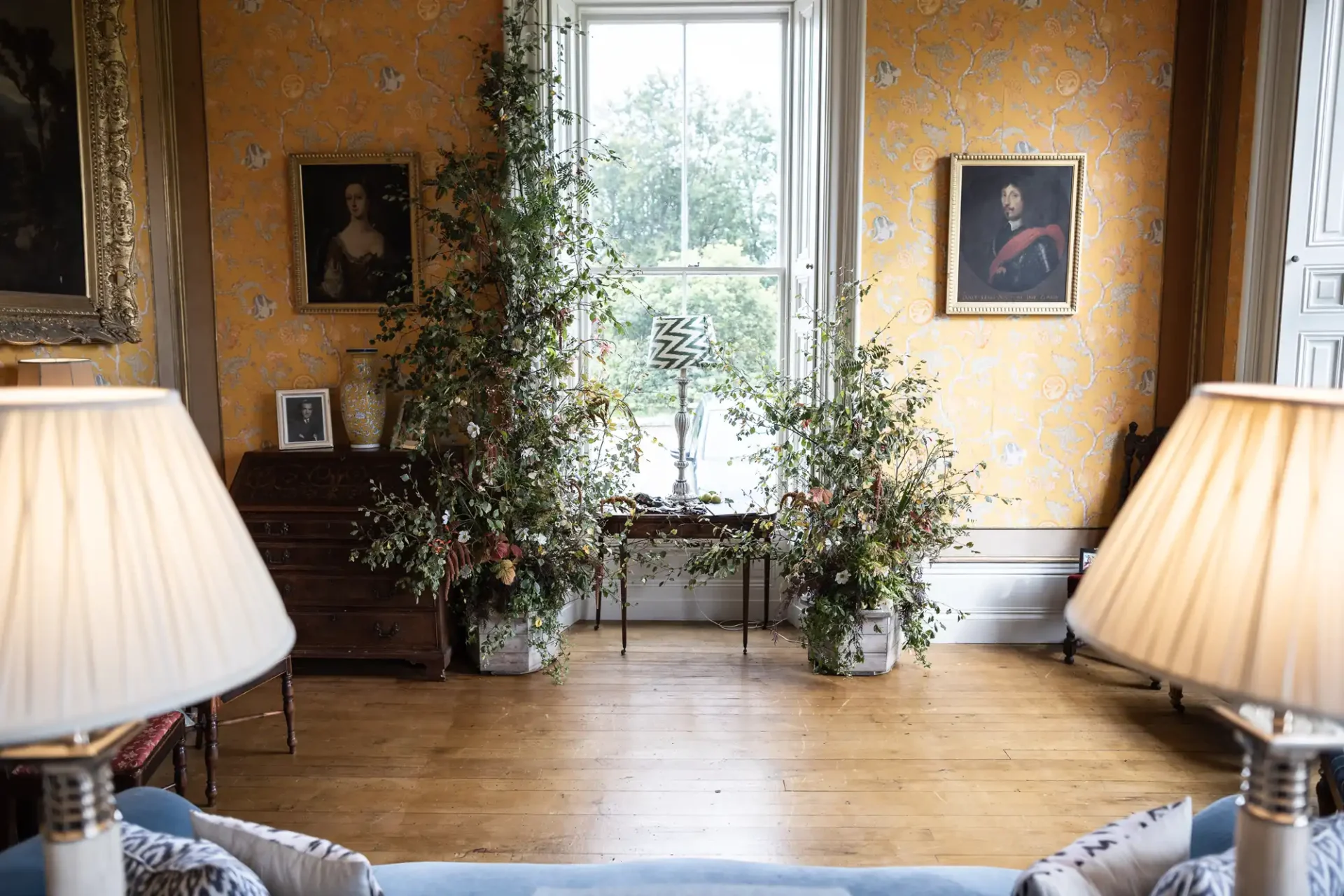 Room with two lamps, a blue sofa, and a window with plants on both sides. Portraits hang on the orange patterned walls. A zigzag sculpture is placed in front of the window.