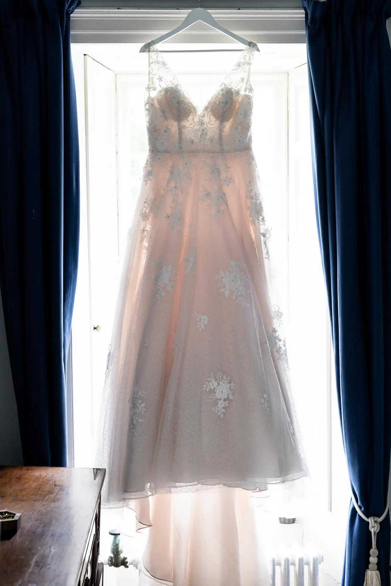 A white lace wedding dress hangs on a hanger in front of a bright window, framed by dark blue curtains.