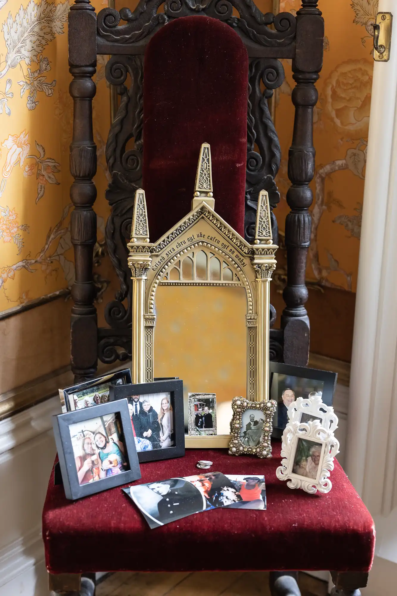 Ornate chair with red velvet seat displays a large decorative mirror and various framed photos, set against a floral wallpaper background.