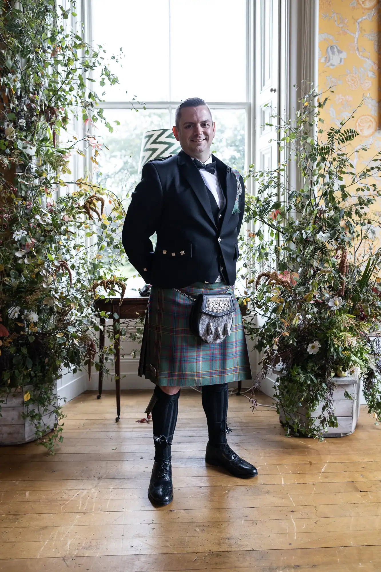 Man in a formal kilt outfit stands in front of a large window framed by plants, in a room with a wooden floor and patterned wallpaper.