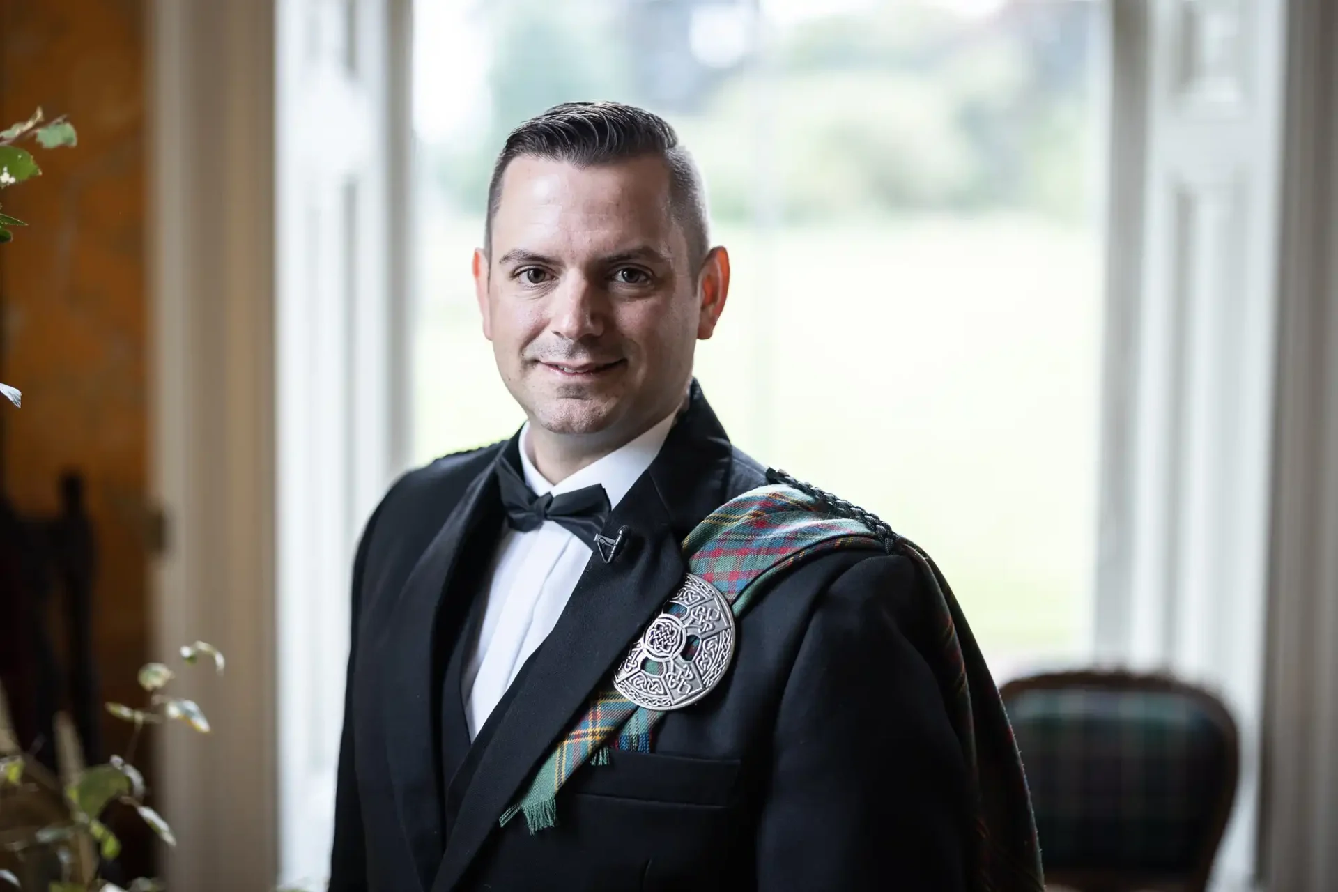 A man in formal Scottish attire with a bow tie and tartan sash stands indoors near a window.