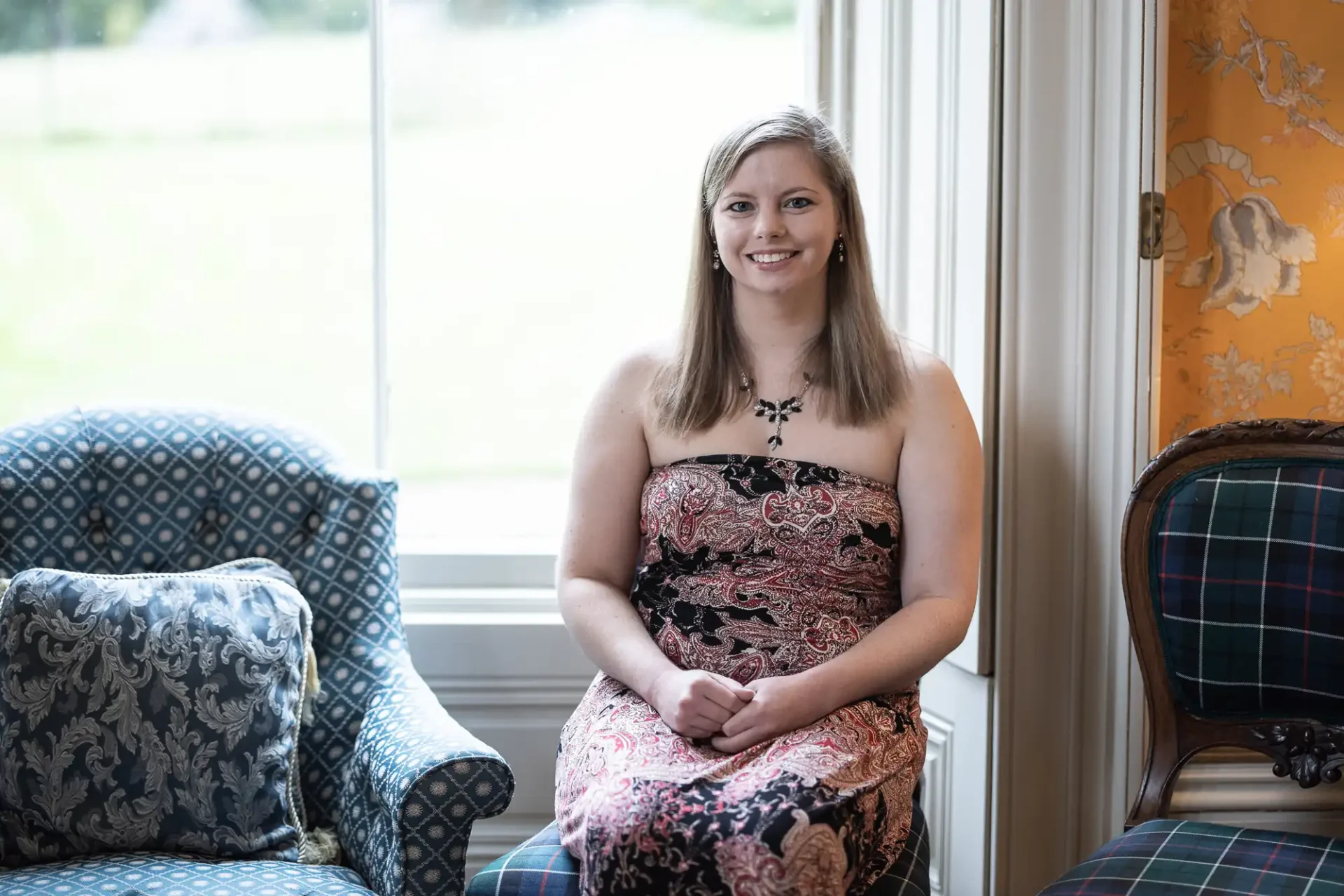 A woman in a patterned dress sits between two chairs near a large window, smiling at the camera.