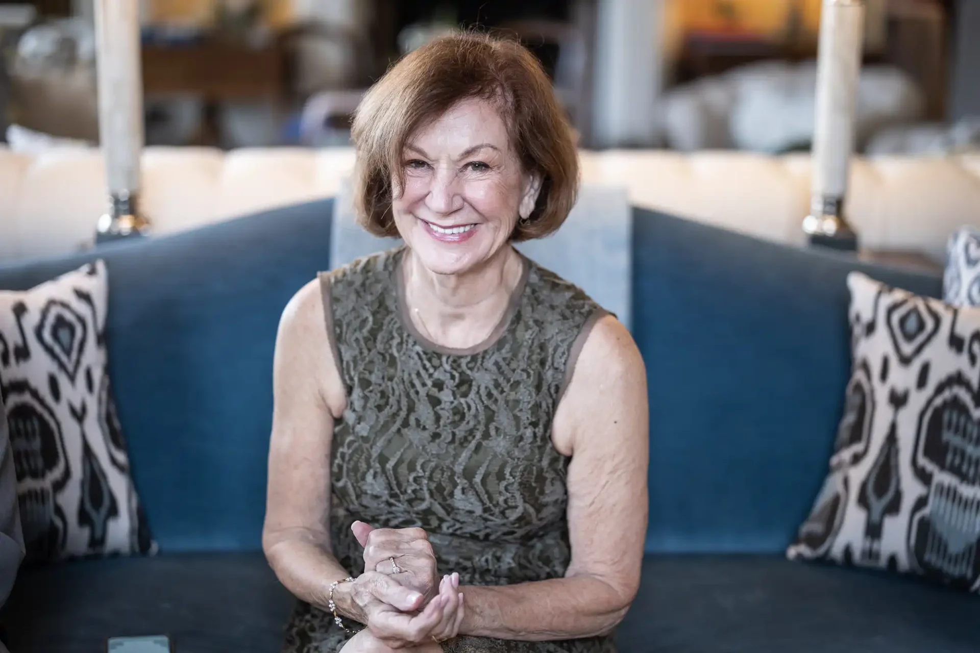 Older woman smiling, seated on a blue couch with patterned pillows, wearing a sleeveless green lace dress.