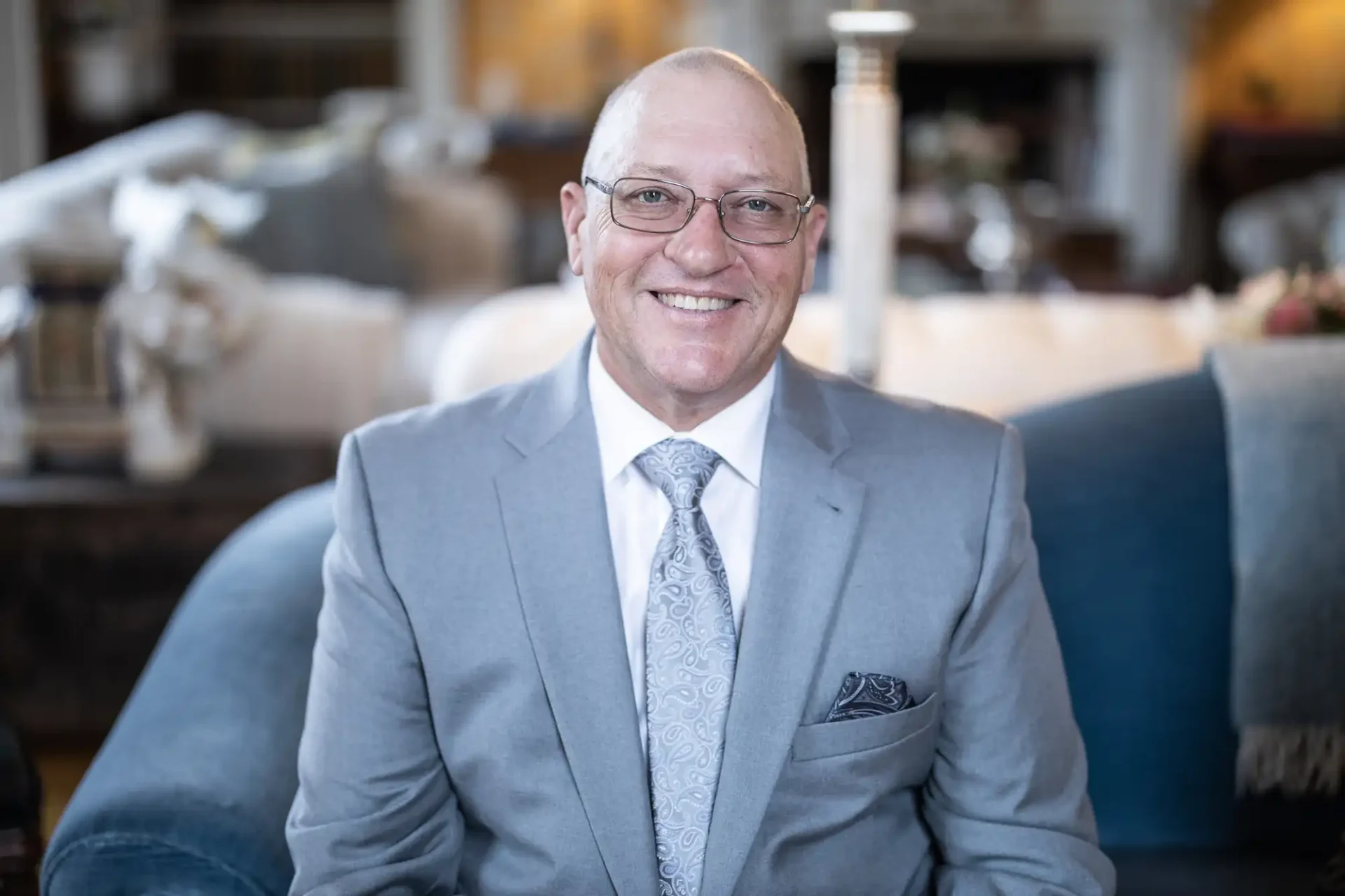 A man in a gray suit and tie, wearing glasses, smiles while seated on a blue couch in a warmly lit room.