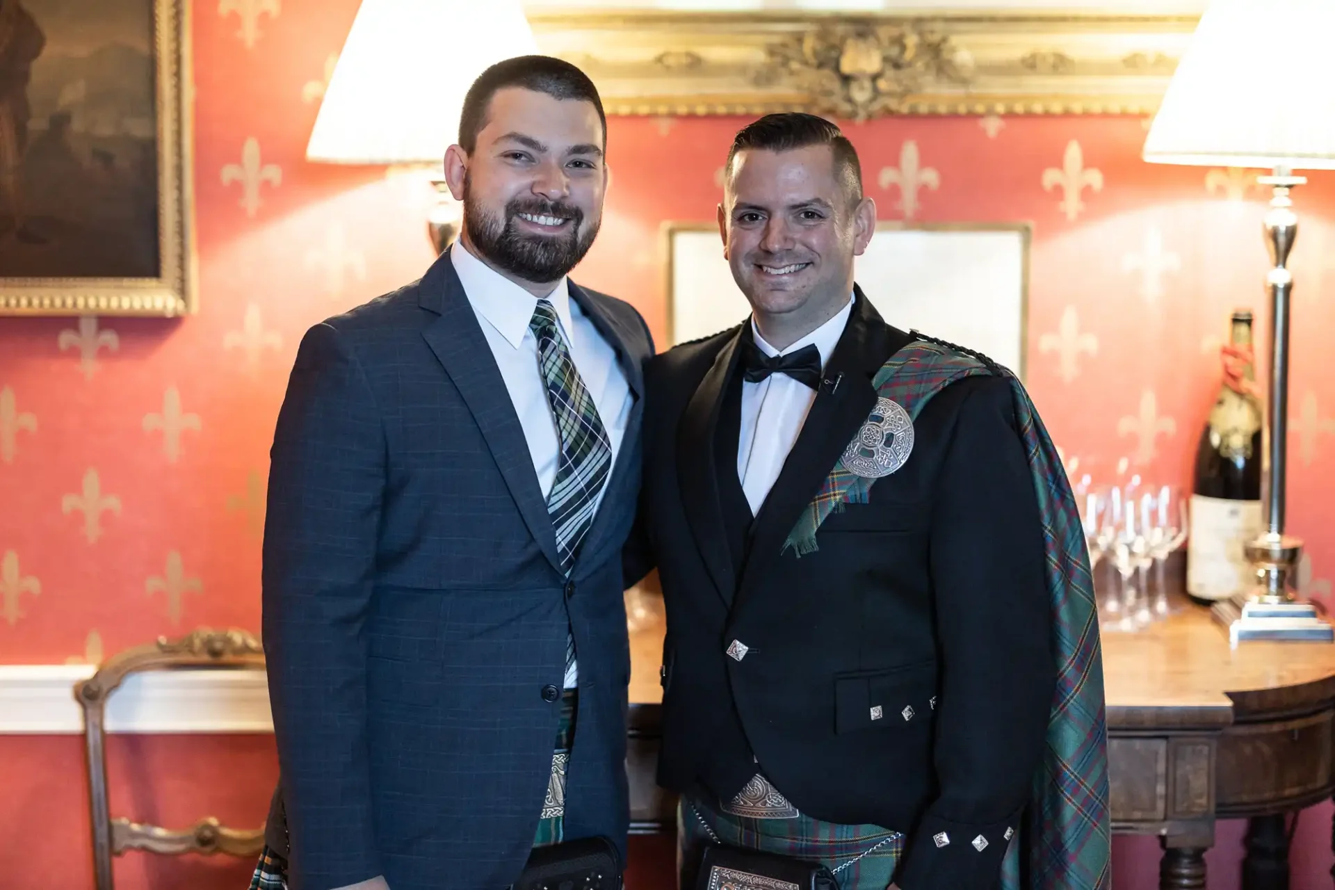 Two men stand side by side. One wears a suit with a tartan tie, and the other wears formal Scottish attire with a kilt. They are in a room with a red patterned wall and lamps.