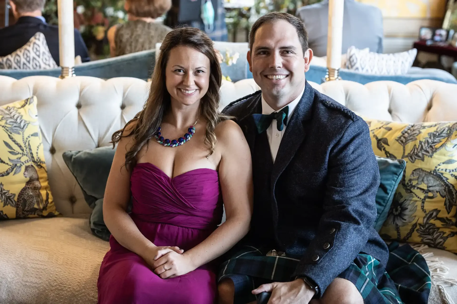 A woman in a pink dress and a man in a suit with a tartan kilt sit on a sofa, smiling at the camera.