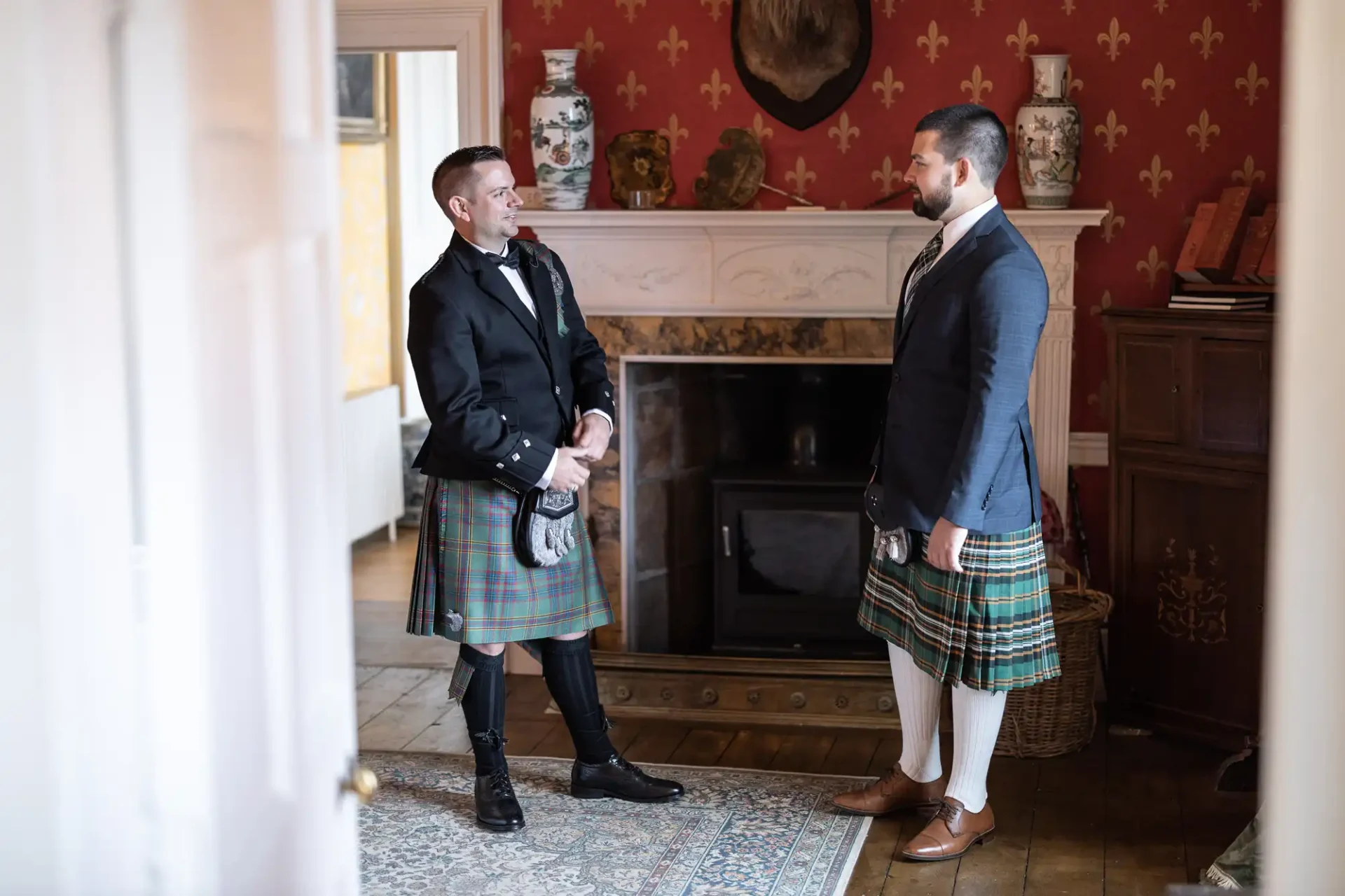 Two men in kilts stand facing each other in a room with red patterned wallpaper and a fireplace.