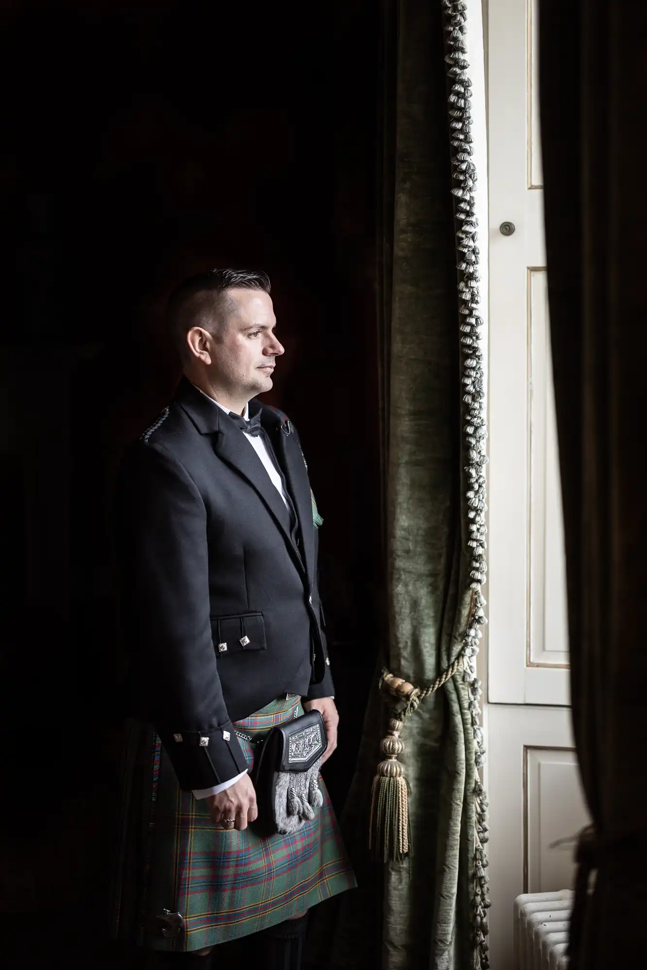 Man in traditional Scottish attire stands by a window, gazing outside.