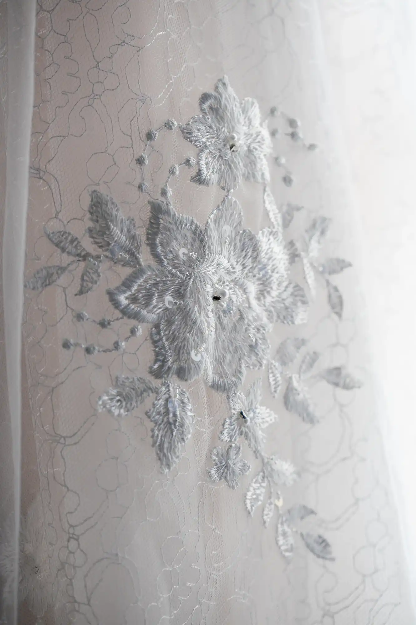 Close-up of intricate floral embroidery on sheer lace fabric, featuring a silver flower pattern with leaves on a light background.