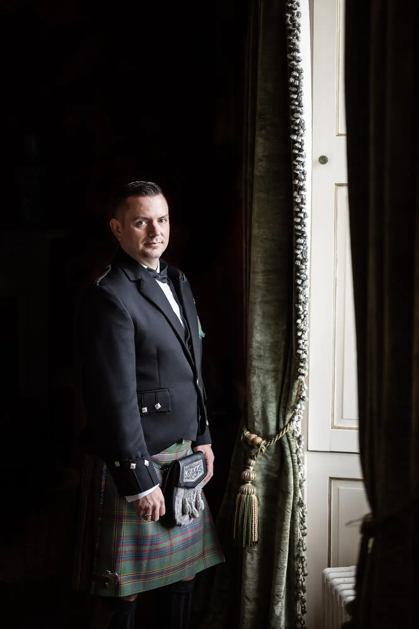 Man in traditional Scottish attire stands by a window with green curtains.