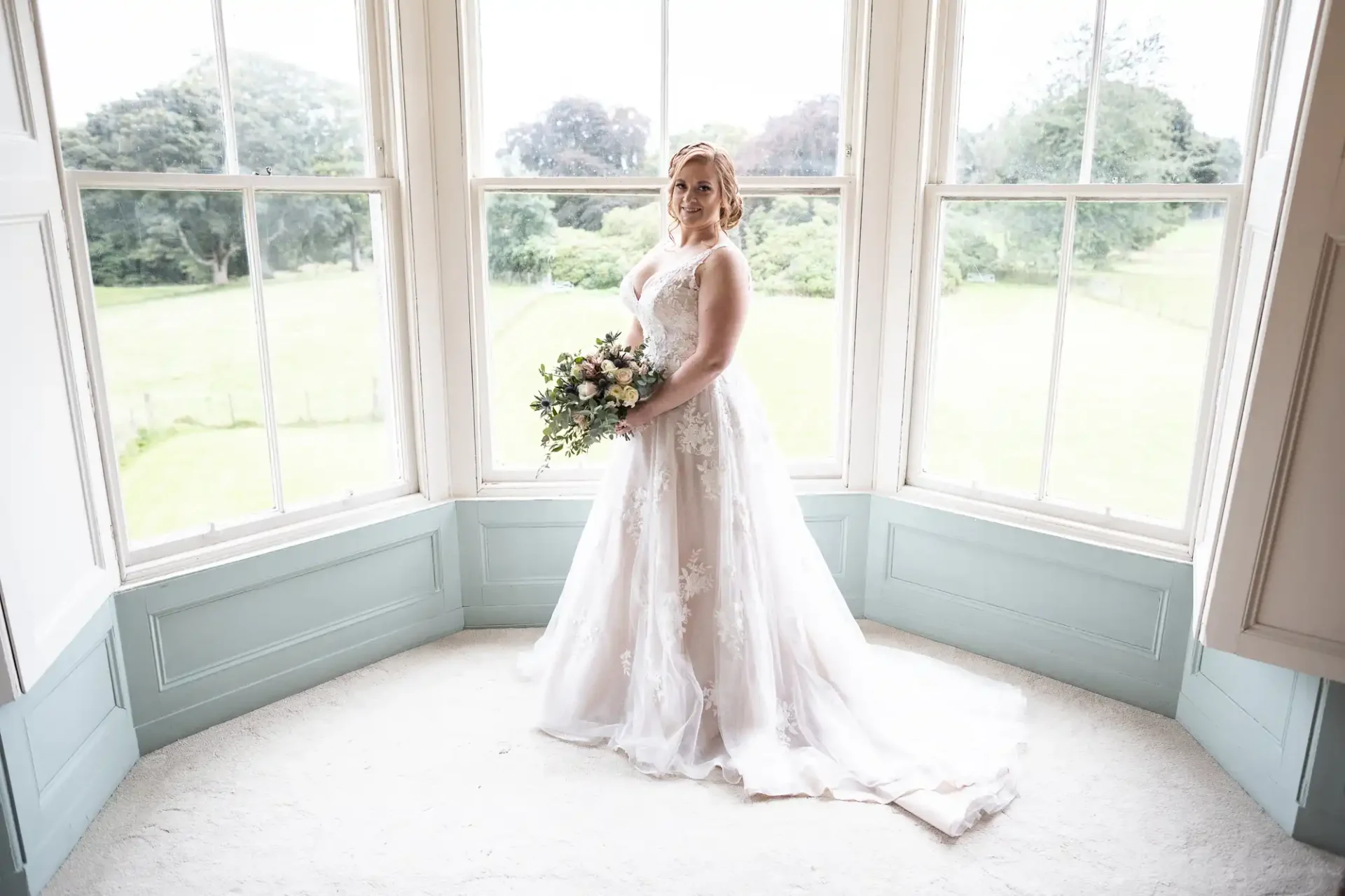 Bride in a white dress holding a bouquet stands by large bay windows, with greenery visible outside.