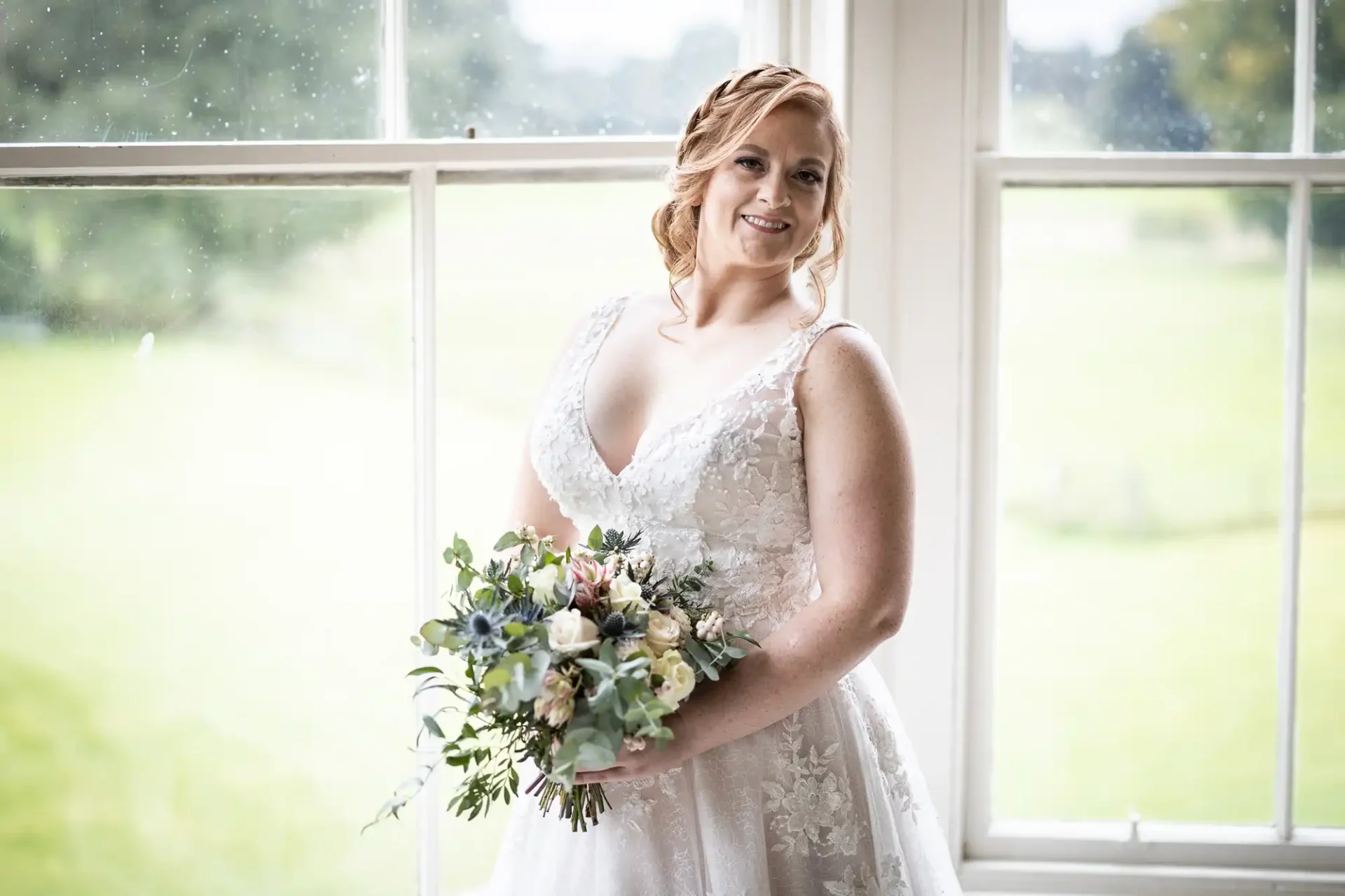 Bride in a white lace gown holds a floral bouquet while standing by a window with a green outdoor view.