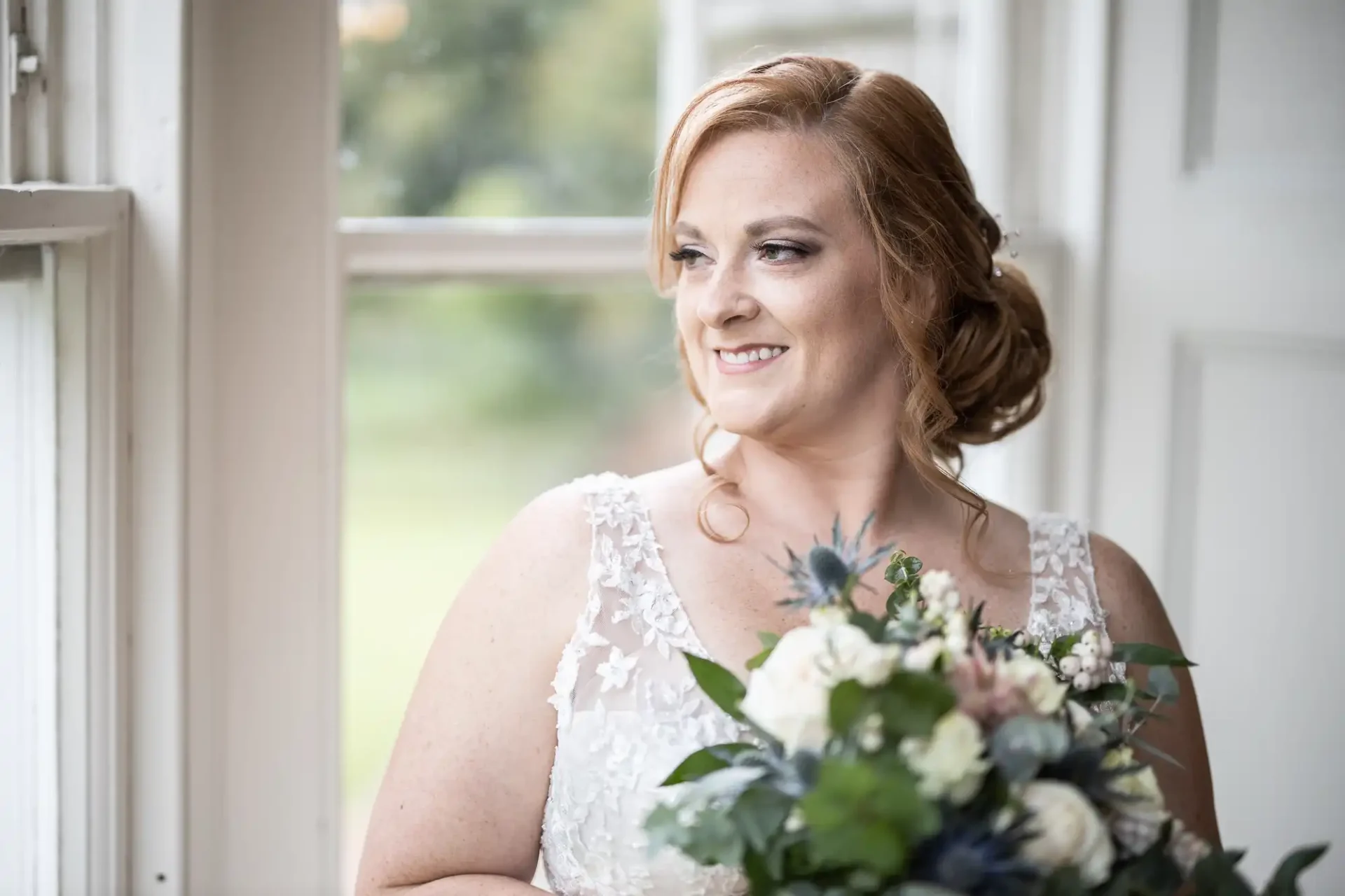 A woman in a white lace dress holds a bouquet, smiling and looking out of a window.