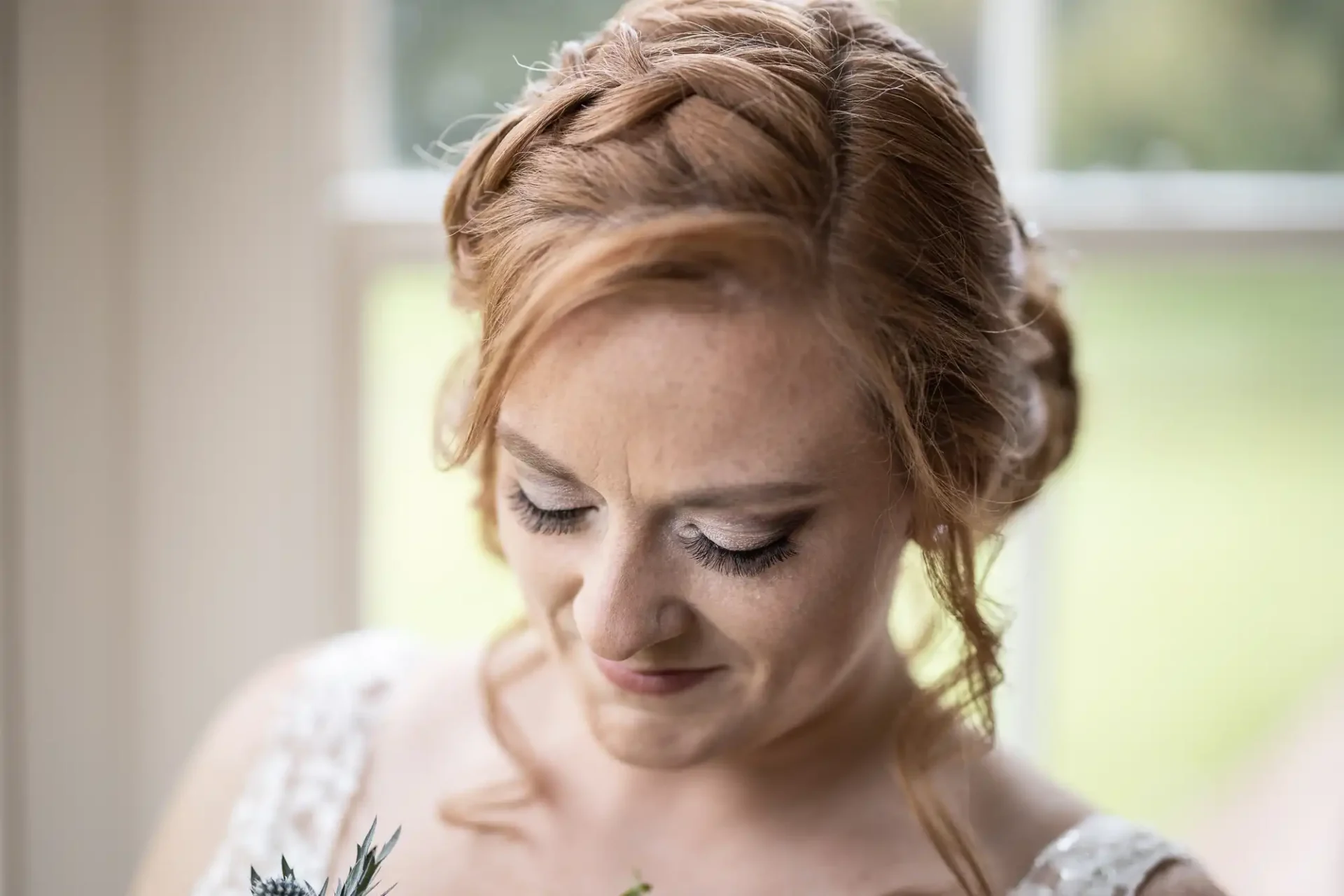 Woman with red hair and a braided updo, wearing a white dress, looks downwards. She is standing in front of a window with a blurred green background.