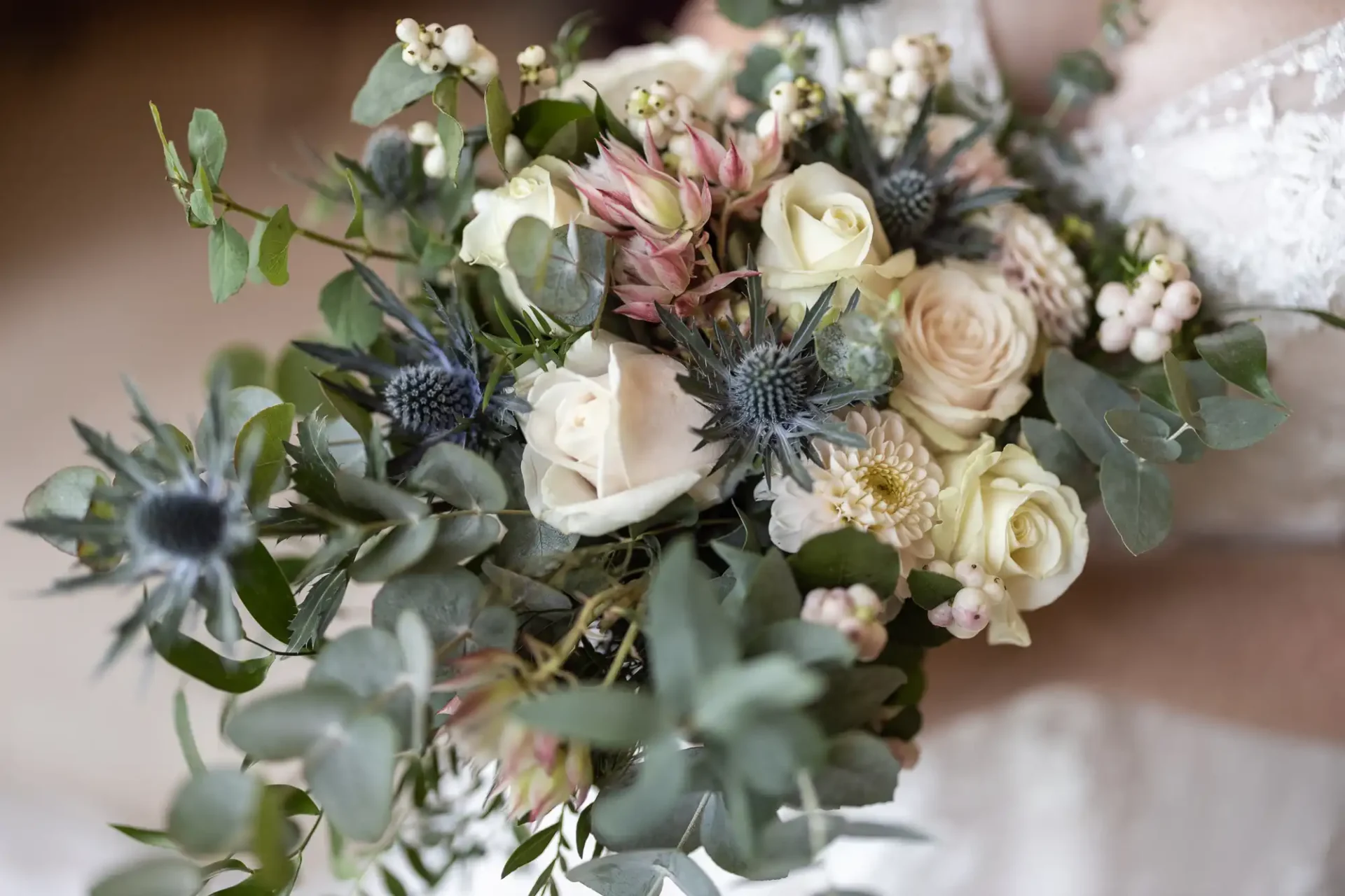 Close-up of a bouquet featuring white and pink roses, eucalyptus leaves, and small thistle flowers, held by a person in a white outfit.