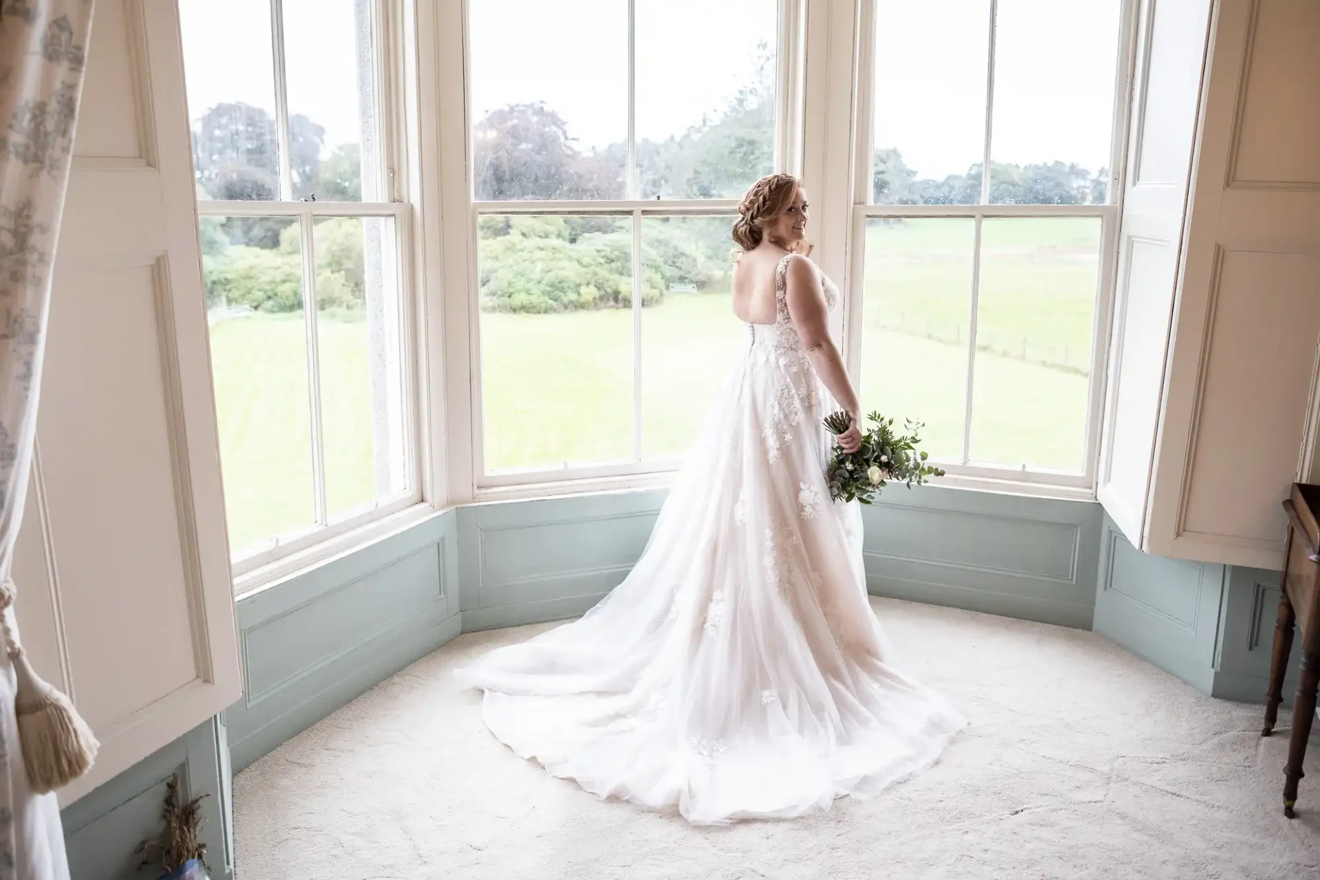 Bride in a white gown stands by large bay windows, holding a bouquet, with a view of a grassy landscape outside.