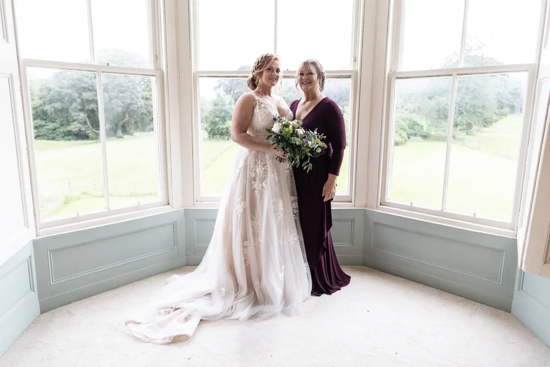 Two women stand side by side in a bay window area. One is in a wedding dress holding a bouquet; the other is in a dark purple gown. They are smiling and looking towards the camera.