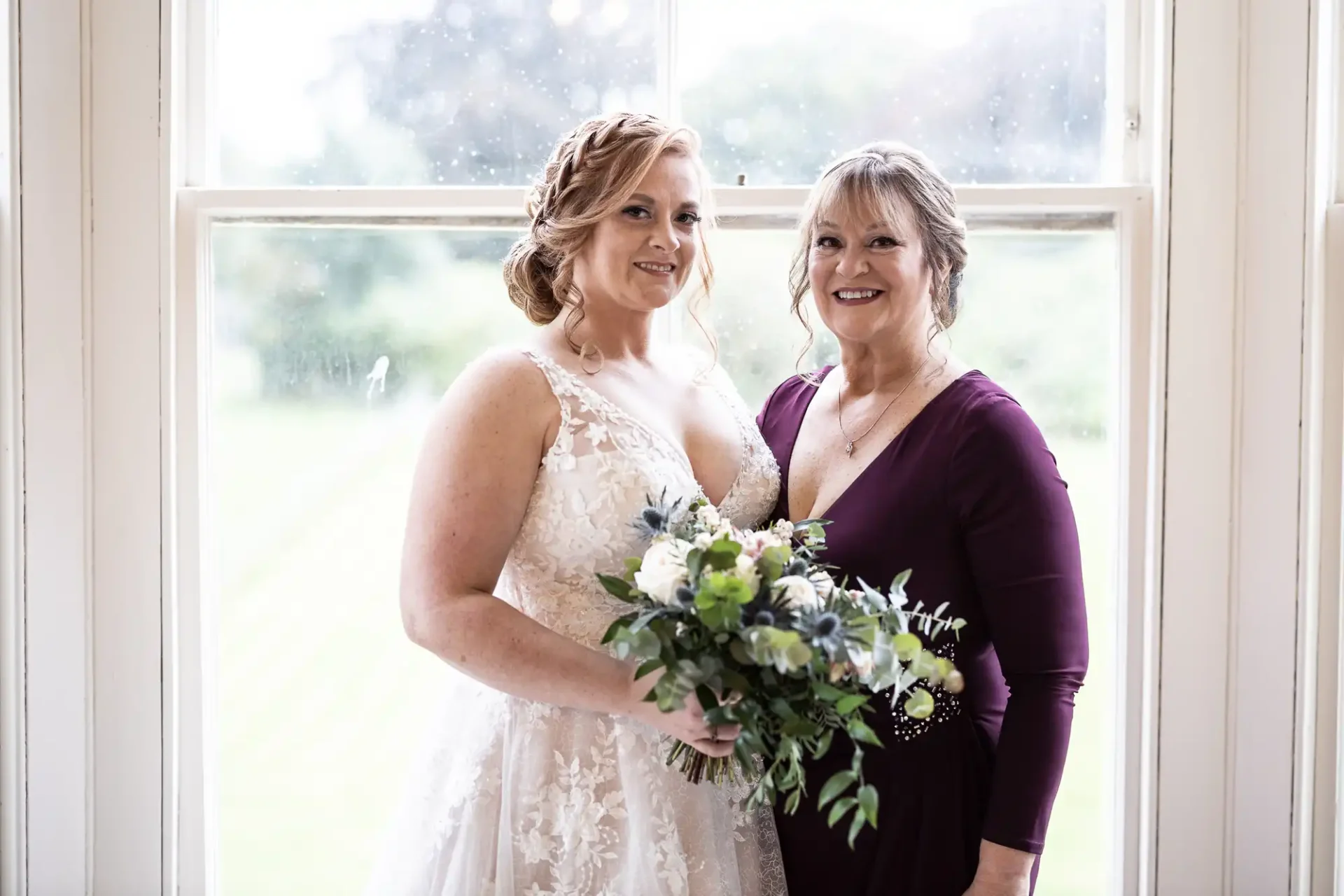 Two women smiling by a window, one in a white wedding dress holding a bouquet, the other in a purple dress.