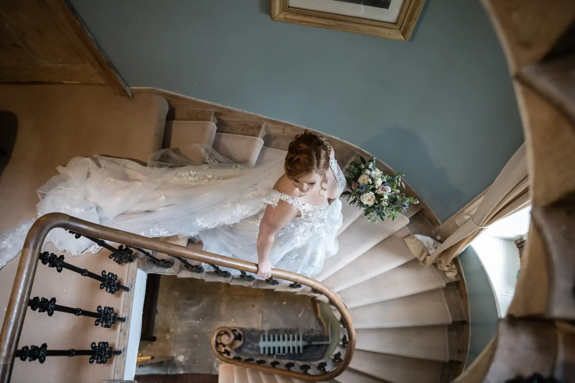 Bride in a lace wedding dress ascends a spiral staircase, holding a bouquet. The scene is viewed from above, capturing the elegant curve of the staircase and her flowing train.