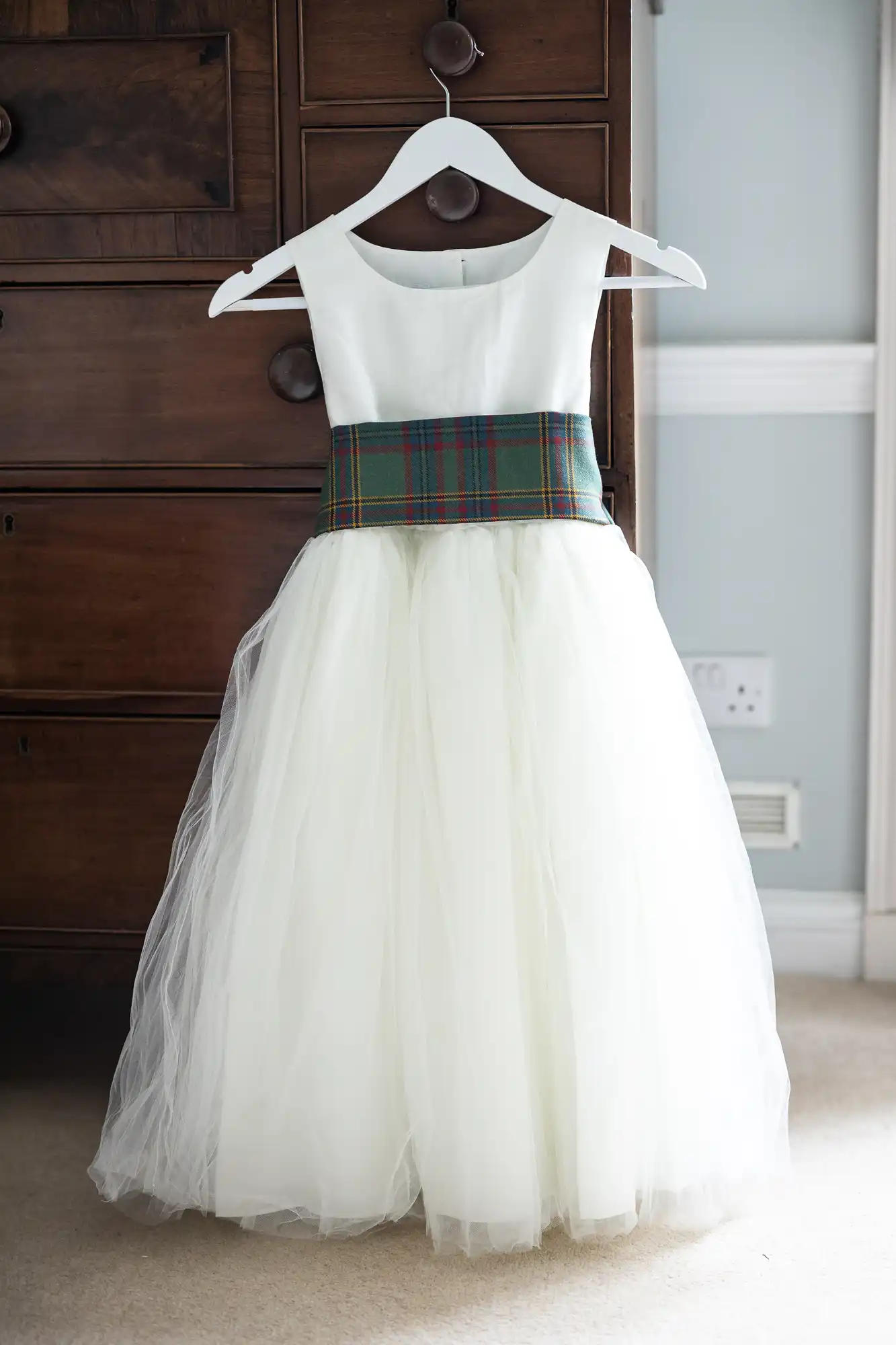 A sleeveless white dress with a tulle skirt and a plaid sash hangs on a white hanger against a dark wooden cabinet.